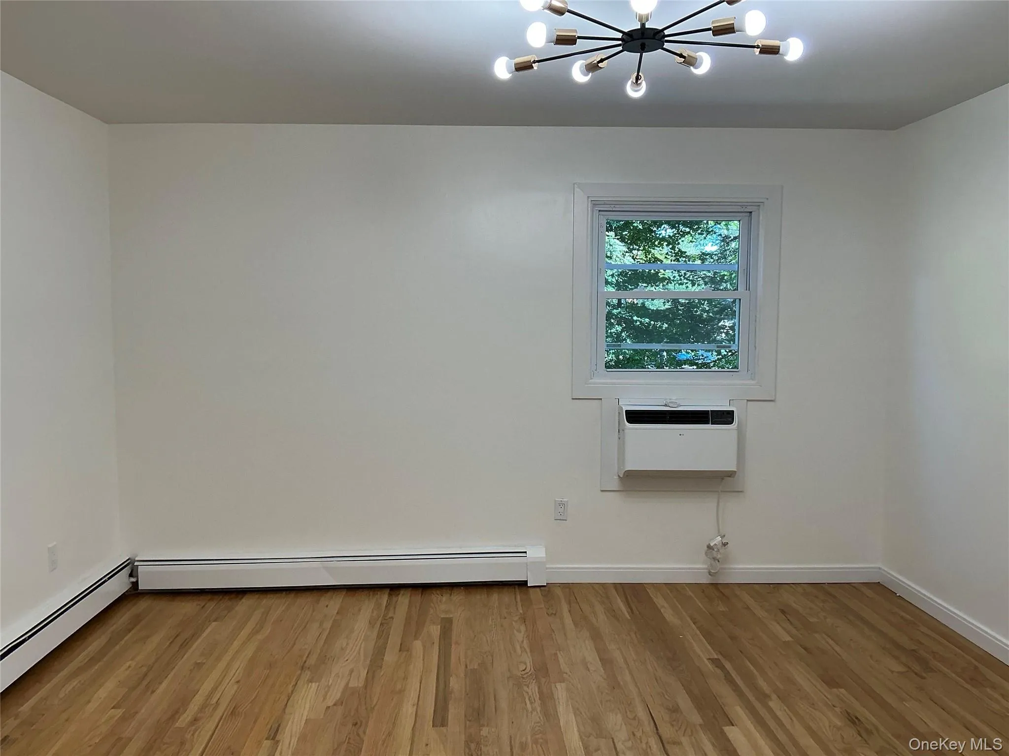 Unfurnished room with a chandelier, light wood-type flooring, a baseboard heating unit, and a wall mounted air conditioner Unfurnished room with a chandelier, light wood-type flooring, a baseboard heating unit, and a wall mounted air conditioner