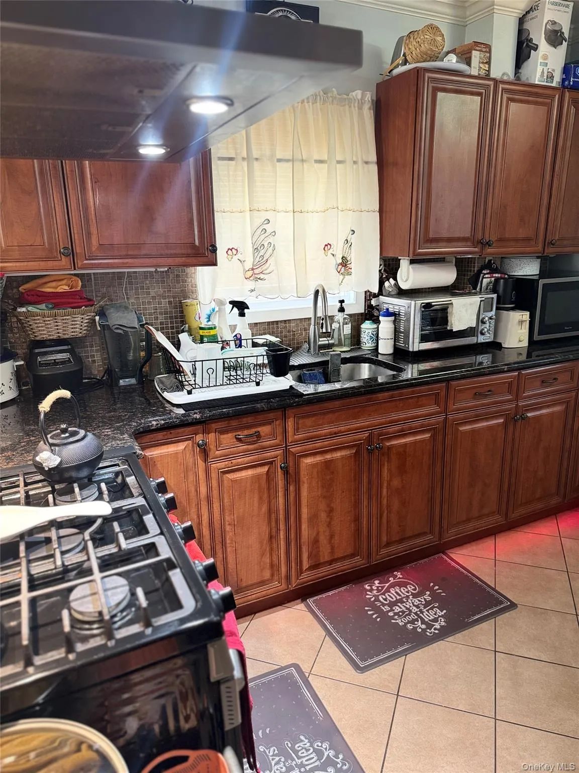 Kitchen featuring under cabinet range hood, gas range oven, backsplash, light tile patterned flooring, and dark stone countertops Kitchen featuring under cabinet range hood, gas range oven, backsplash, light tile patterned flooring, and dark stone countertops