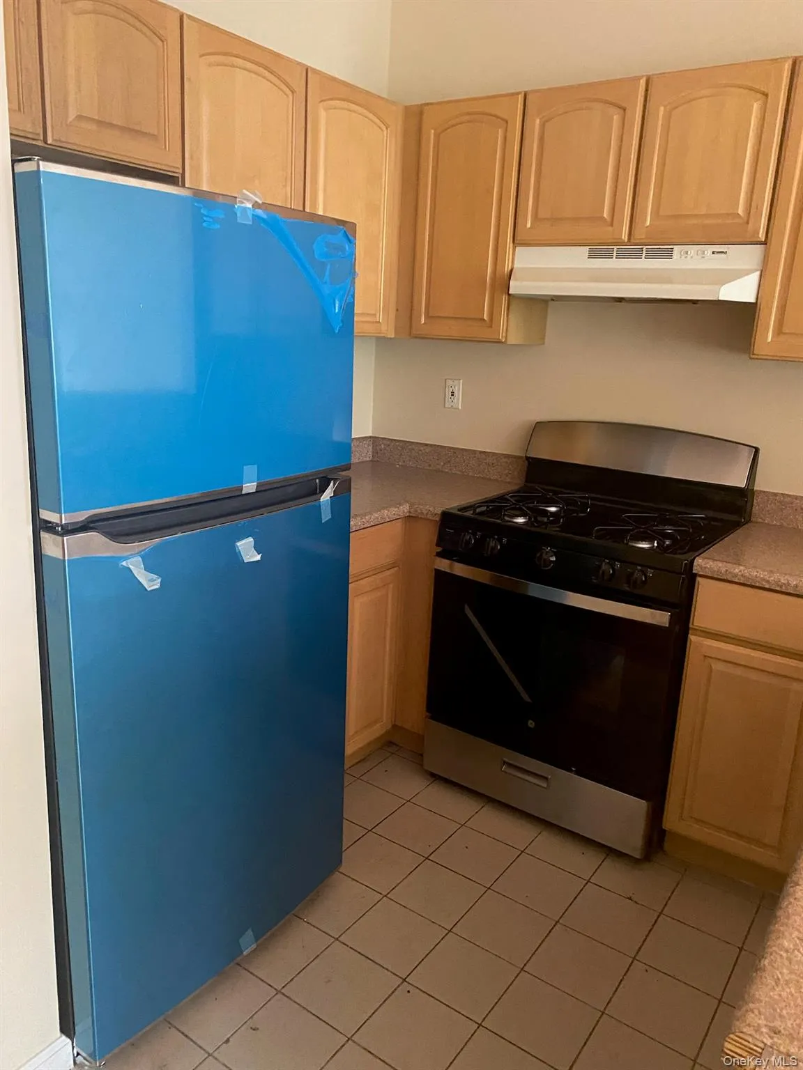 Kitchen with gas stove, freestanding refrigerator, light tile patterned floors, under cabinet range hood, and light brown cabinetry Kitchen with gas stove, freestanding refrigerator, light tile patterned floors, under cabinet range hood, and light brown cabinetry