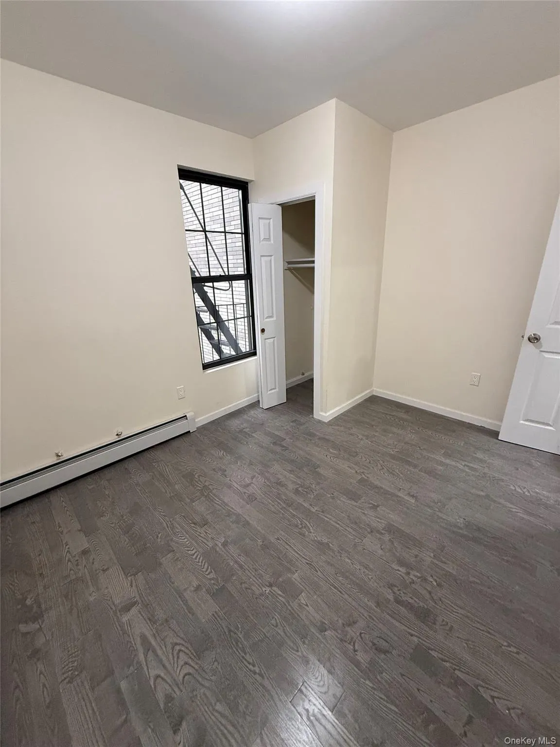 Unfurnished bedroom with a baseboard radiator, dark wood-style floors, and a spacious closet Unfurnished bedroom with a baseboard radiator, dark wood-style floors, and a spacious closet