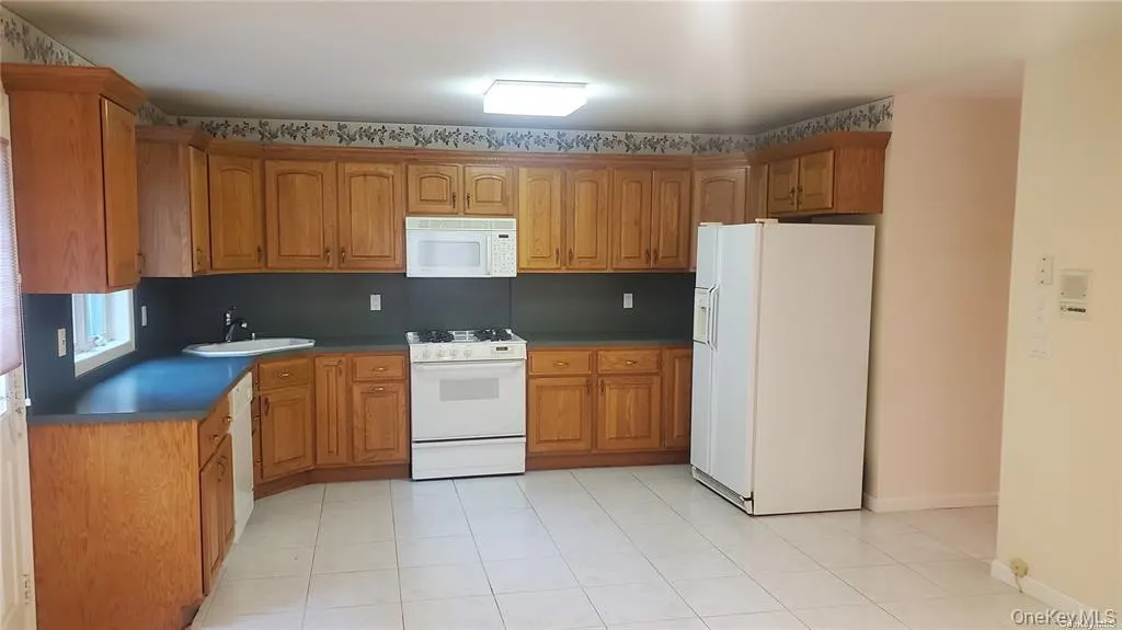 Kitchen featuring white appliances, brown cabinetry, light tile patterned floors, and dark countertops Kitchen featuring white appliances, brown cabinetry, light tile patterned floors, and dark countertops