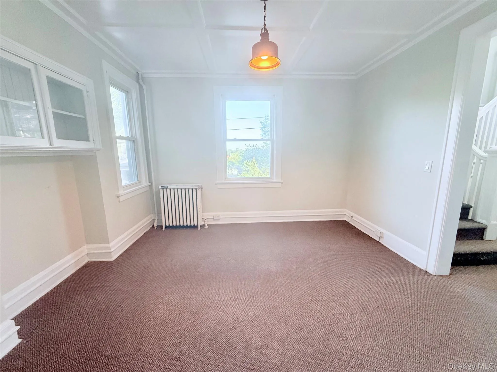 Empty room featuring radiator heating unit, coffered ceiling, carpet, stairs, and ornamental molding Empty room featuring radiator heating unit, coffered ceiling, carpet, stairs, and ornamental molding