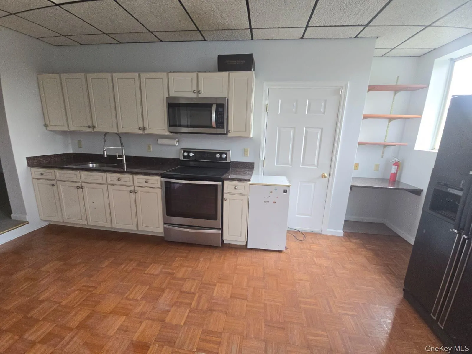 Kitchen with stainless steel appliances, a paneled ceiling, cream cabinetry, and open shelves Kitchen with stainless steel appliances, a paneled ceiling, cream cabinetry, and open shelves
