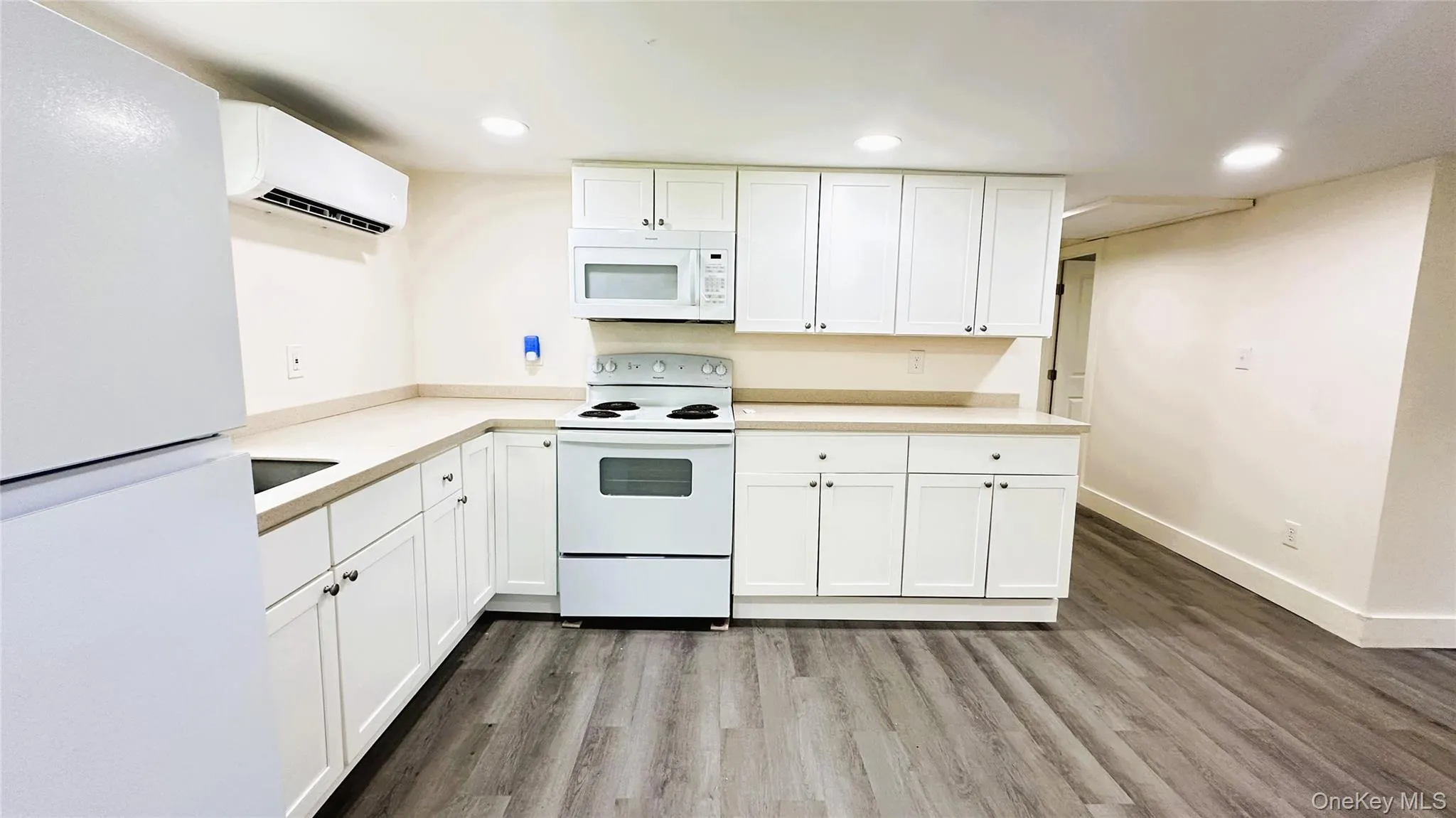 Kitchen with white appliances, an AC wall unit, light vinyl  flooring, white cabinetry, and recessed lighting Kitchen with white appliances, an AC wall unit, light vinyl  flooring, white cabinetry, and recessed lighting