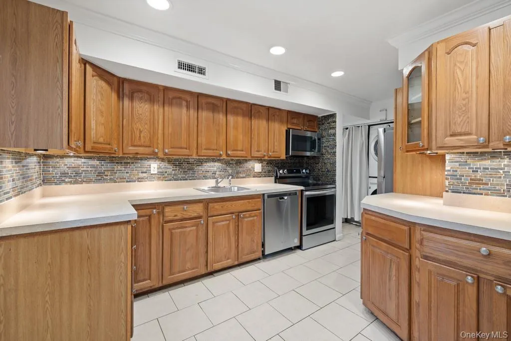 Kitchen featuring decorative backsplash, brown cabinets, light countertops, appliances with stainless steel finishes, and stacked washer / dryer Kitchen featuring decorative backsplash, brown cabinets, light countertops, appliances with stainless steel finishes, and stacked washer / dryer