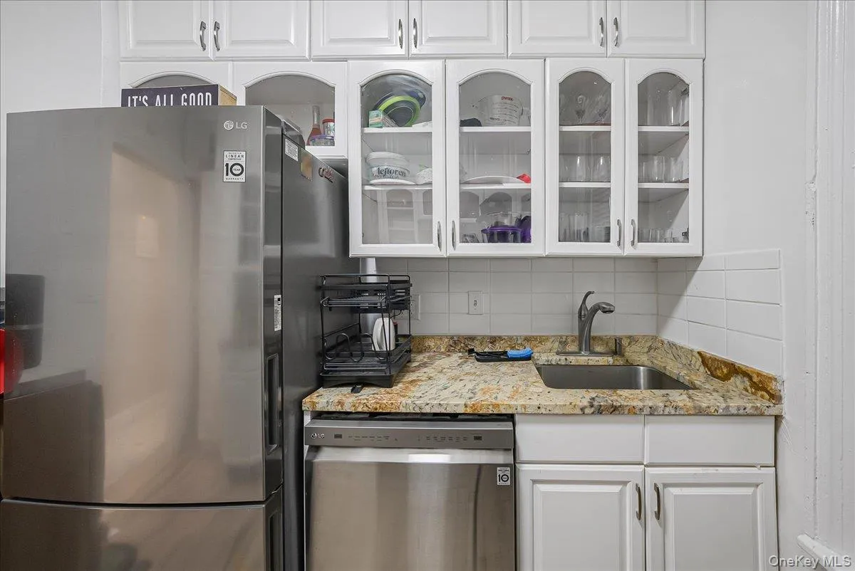 Kitchen with white cabinetry, decorative backsplash, stainless steel appliances, glass insert cabinets, and light stone counters Kitchen with white cabinetry, decorative backsplash, stainless steel appliances, glass insert cabinets, and light stone counters