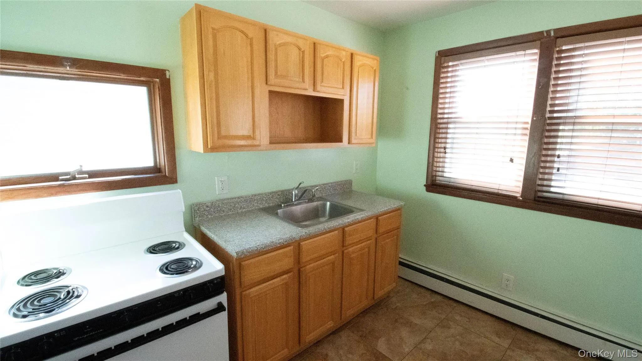 Kitchen featuring white electric range oven, a baseboard heating unit, light brown cabinetry, light countertops, and dark tile patterned flooring Kitchen featuring white electric range oven, a baseboard heating unit, light brown cabinetry, light countertops, and dark tile patterned flooring