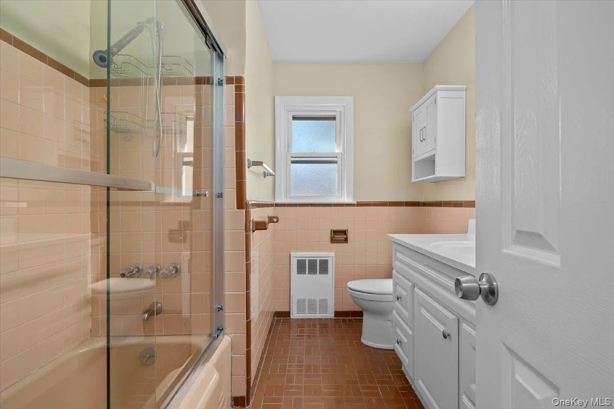 Bathroom with vanity, shower / bath combination with glass door, a wainscoted wall, tile walls, and radiator heating unit Bathroom with vanity, shower / bath combination with glass door, a wainscoted wall, tile walls, and radiator heating unit