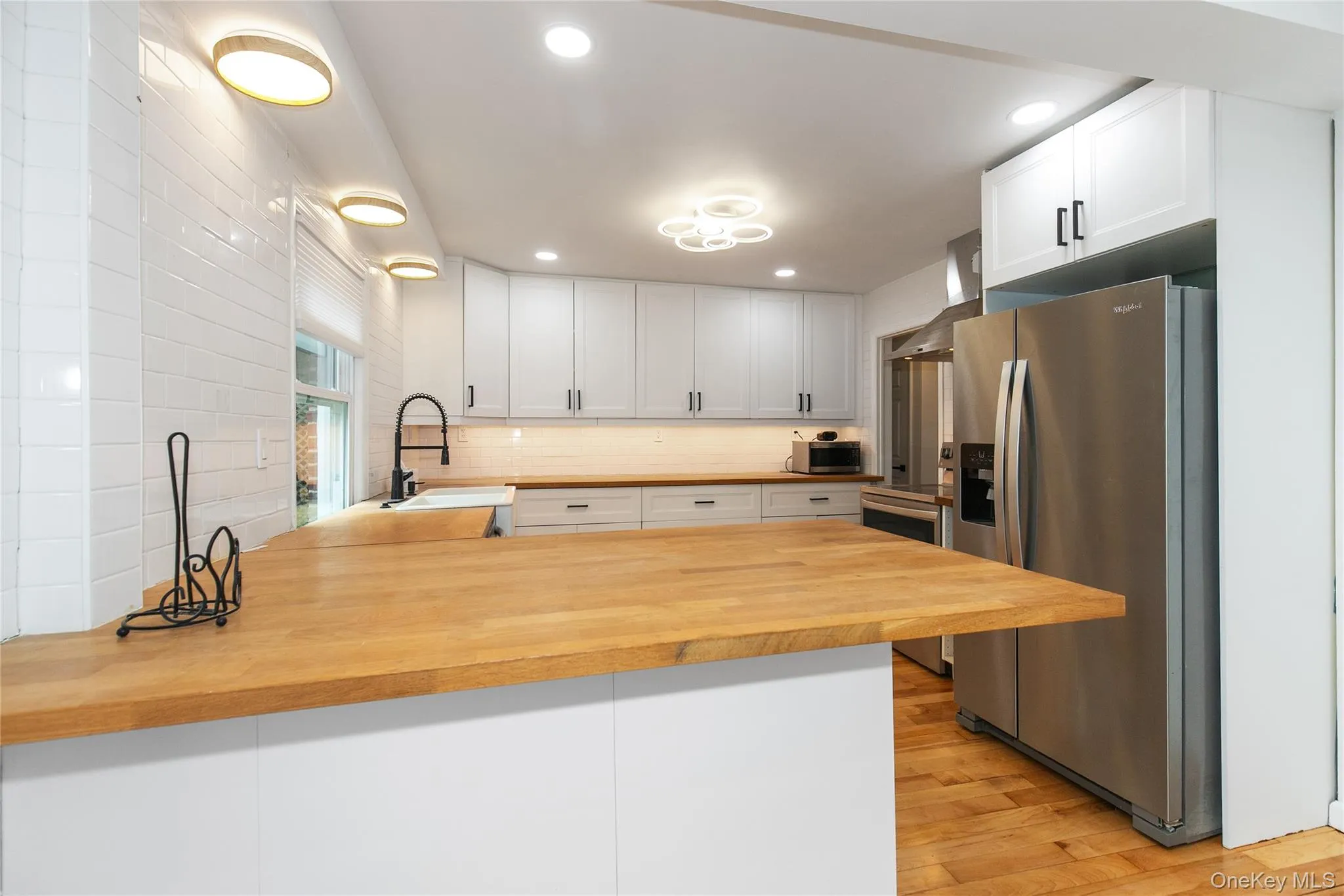 Kitchen featuring a peninsula, stainless steel fridge, white cabinets, wall chimney range hood, and light wood-style floors Kitchen featuring a peninsula, stainless steel fridge, white cabinets, wall chimney range hood, and light wood-style floors