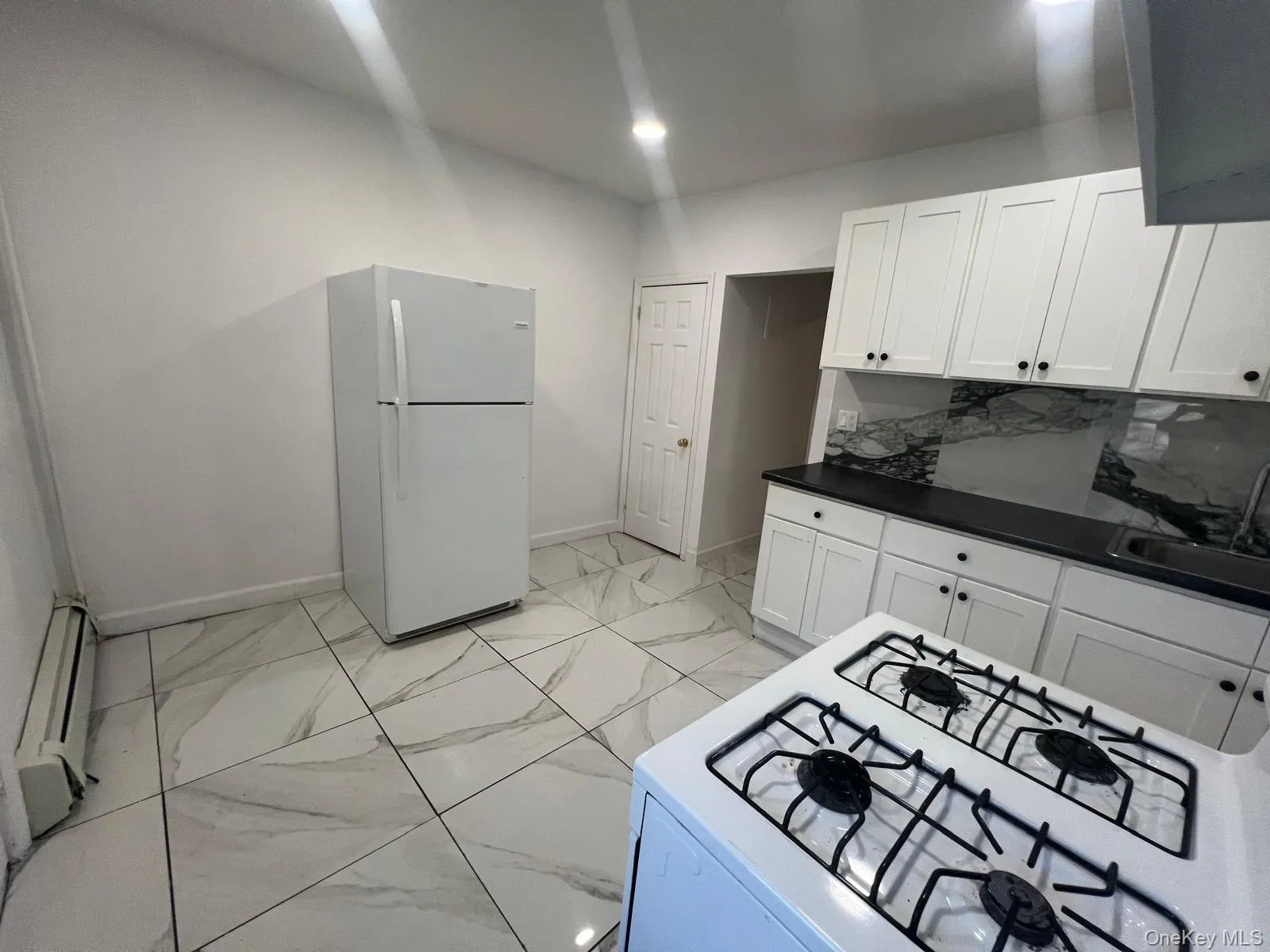 Kitchen featuring dark countertops, white appliances, light marble finish flooring, baseboard heating, and white cabinets Kitchen featuring dark countertops, white appliances, light marble finish flooring, baseboard heating, and white cabinets