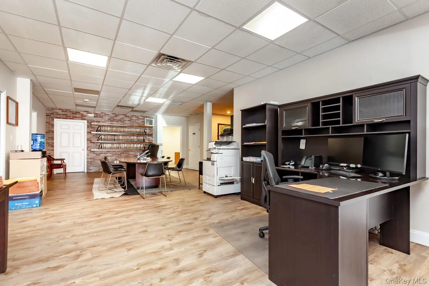 Office area with a drop ceiling, light wood-style flooring, and brick wall Office area with a drop ceiling, light wood-style flooring, and brick wall