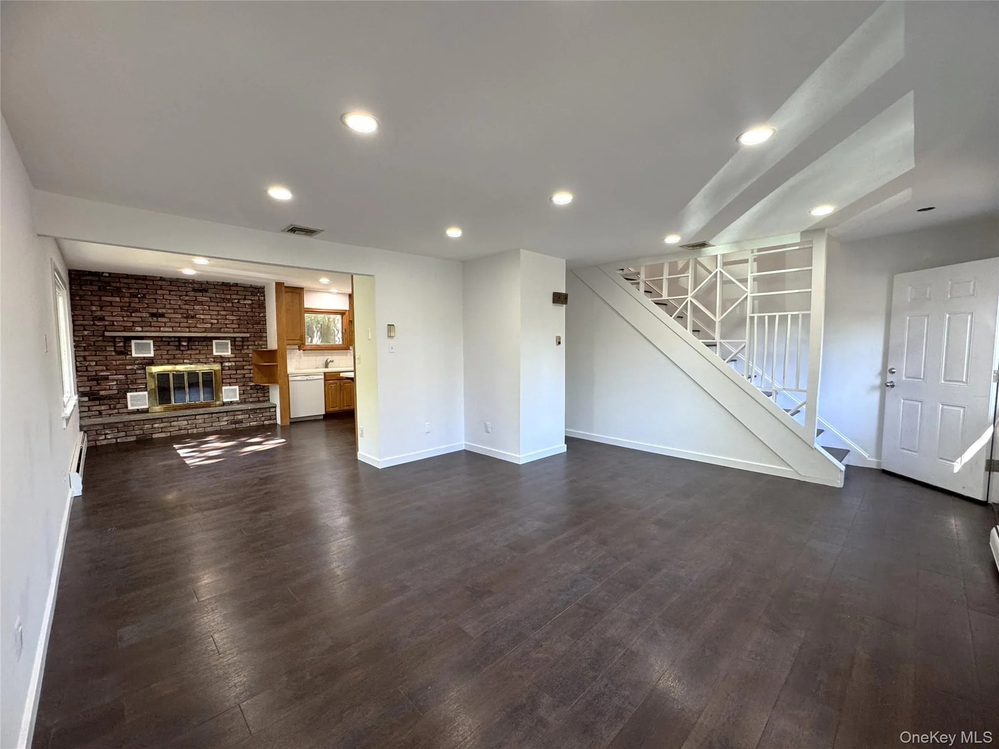 Unfurnished living room featuring a brick fireplace, stairway, dark wood-type flooring, recessed lighting, and baseboard heating Unfurnished living room featuring a brick fireplace, stairway, dark wood-type flooring, recessed lighting, and baseboard heating