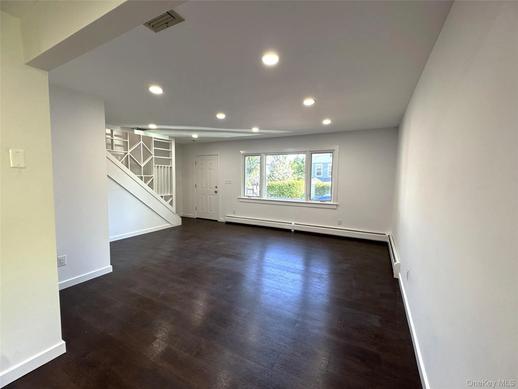 Unfurnished living room featuring dark wood-style flooring, recessed lighting, a baseboard heating unit, and stairs Unfurnished living room featuring dark wood-style flooring, recessed lighting, a baseboard heating unit, and stairs