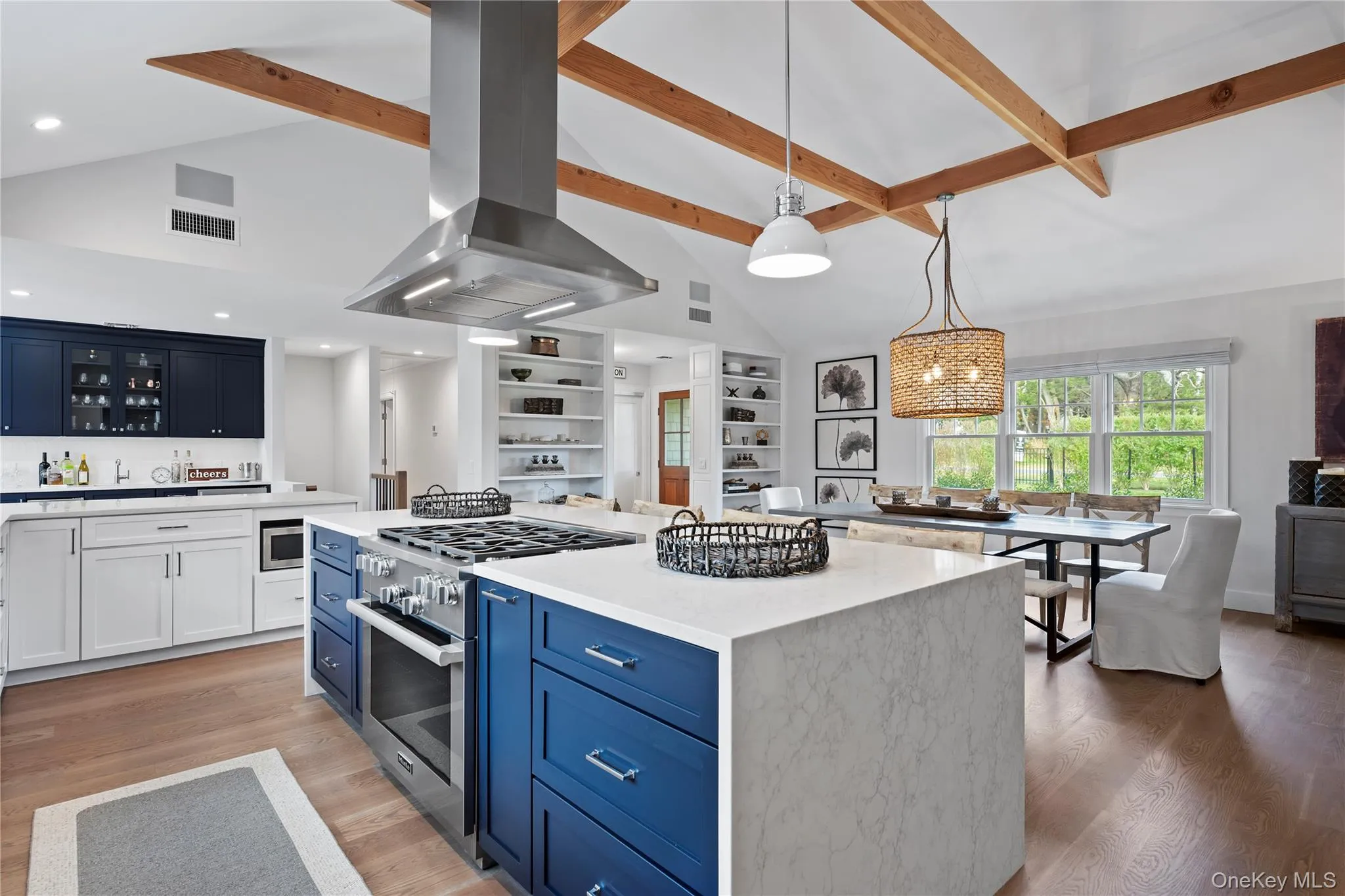 Kitchen featuring blue cabinetry, range hood, appliances with stainless steel finishes, hanging light fixtures, and light stone counters