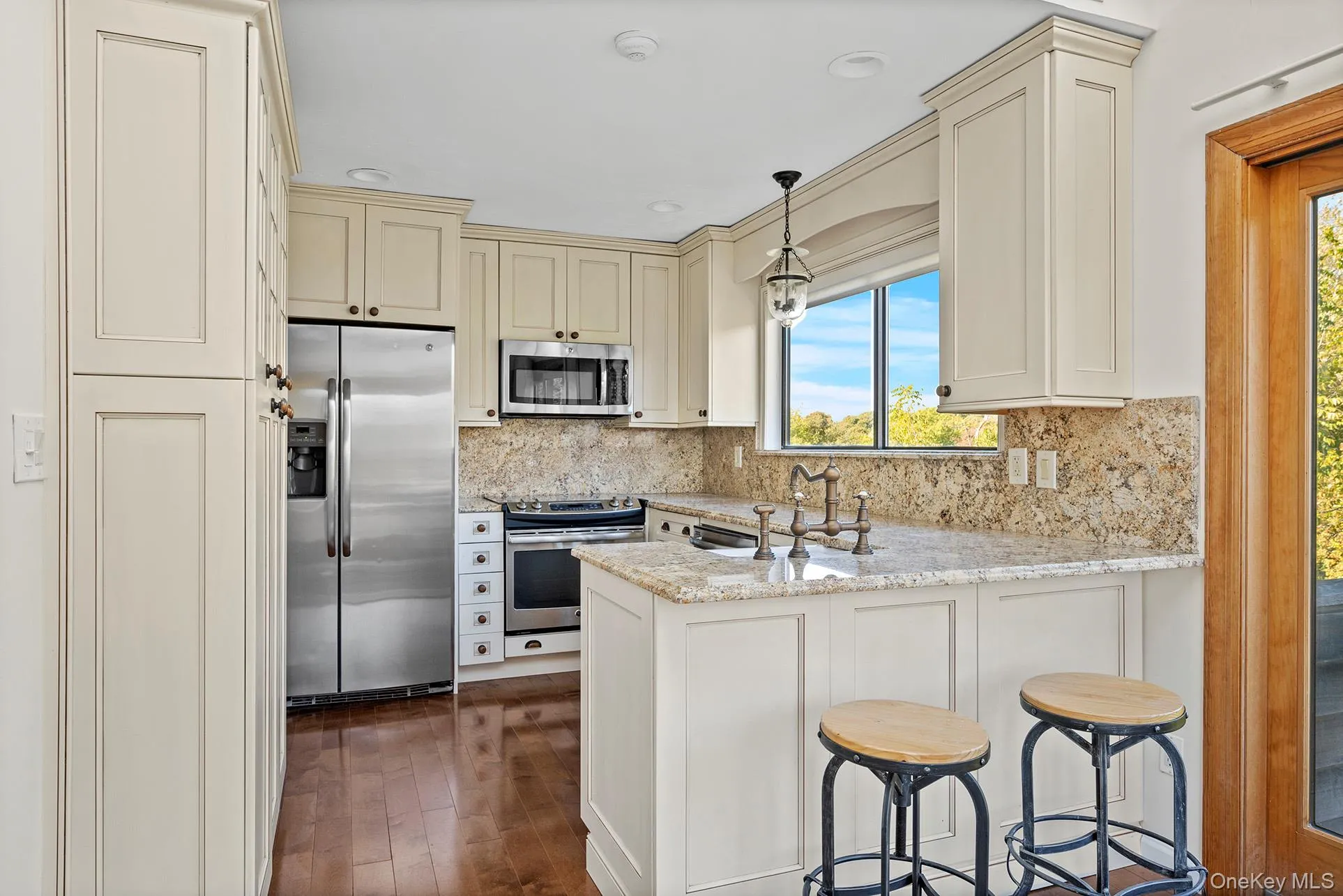 Kitchen with cream cabinetry, decorative light fixtures, stainless steel appliances, a peninsula, and light stone counters Kitchen with cream cabinetry, decorative light fixtures, stainless steel appliances, a peninsula, and light stone counters