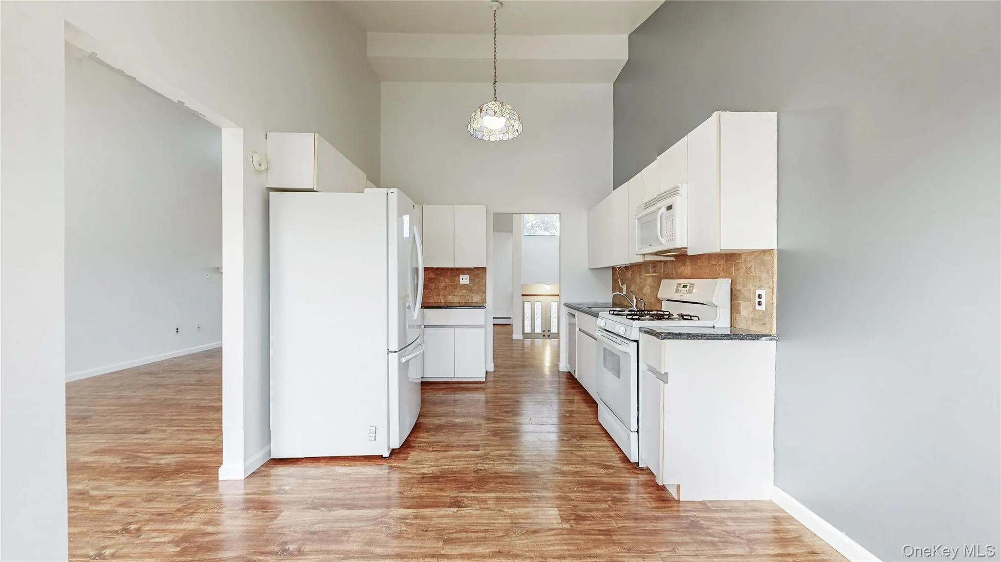 Kitchen with a high ceiling, white appliances, backsplash, light wood finished floors, and decorative light fixtures Kitchen with a high ceiling, white appliances, backsplash, light wood finished floors, and decorative light fixtures