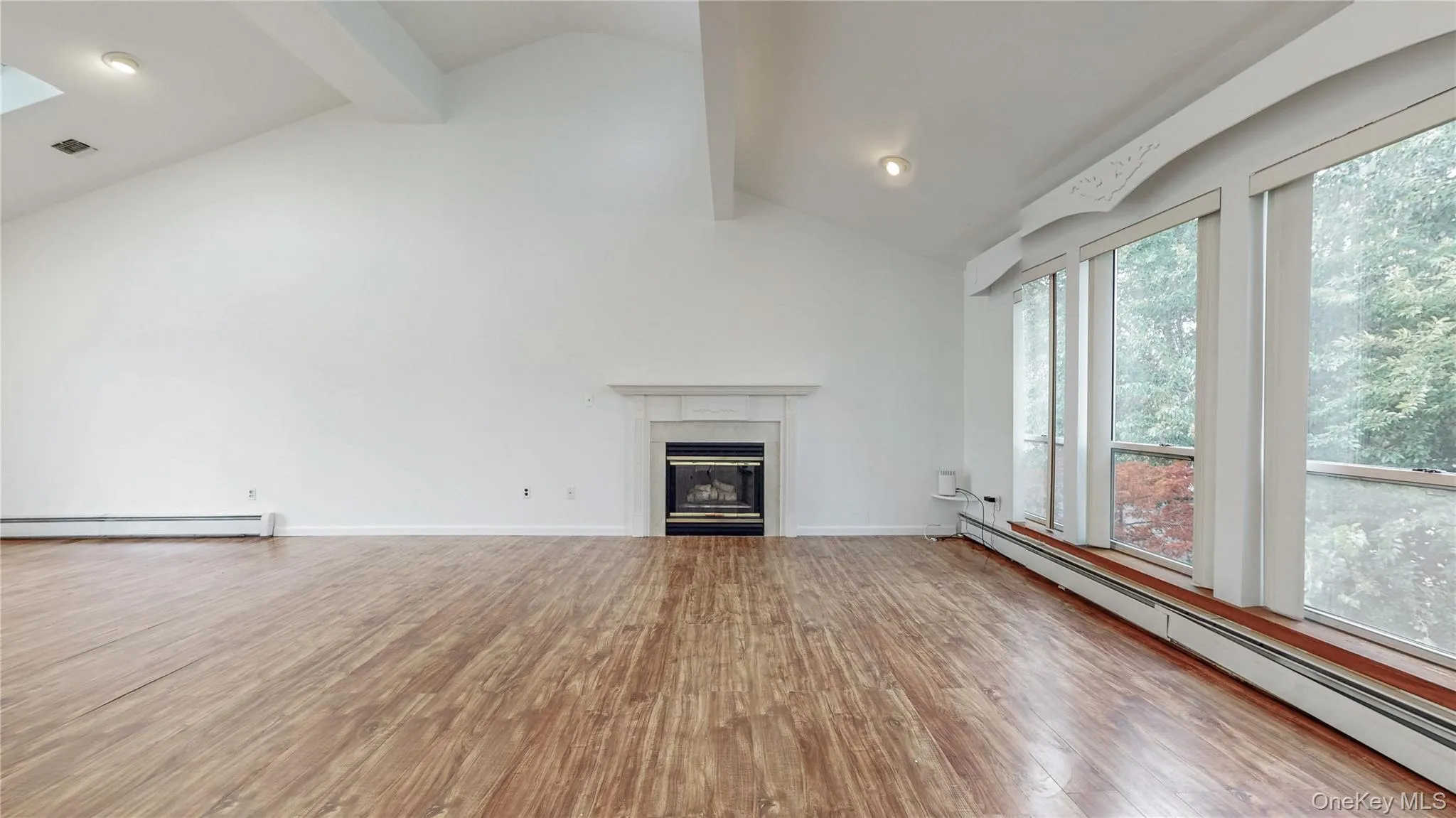 Unfurnished living room with beam ceiling, baseboard heating, light wood-type flooring, a glass covered fireplace, and high vaulted ceiling Unfurnished living room with beam ceiling, baseboard heating, light wood-type flooring, a glass covered fireplace, and high vaulted ceiling