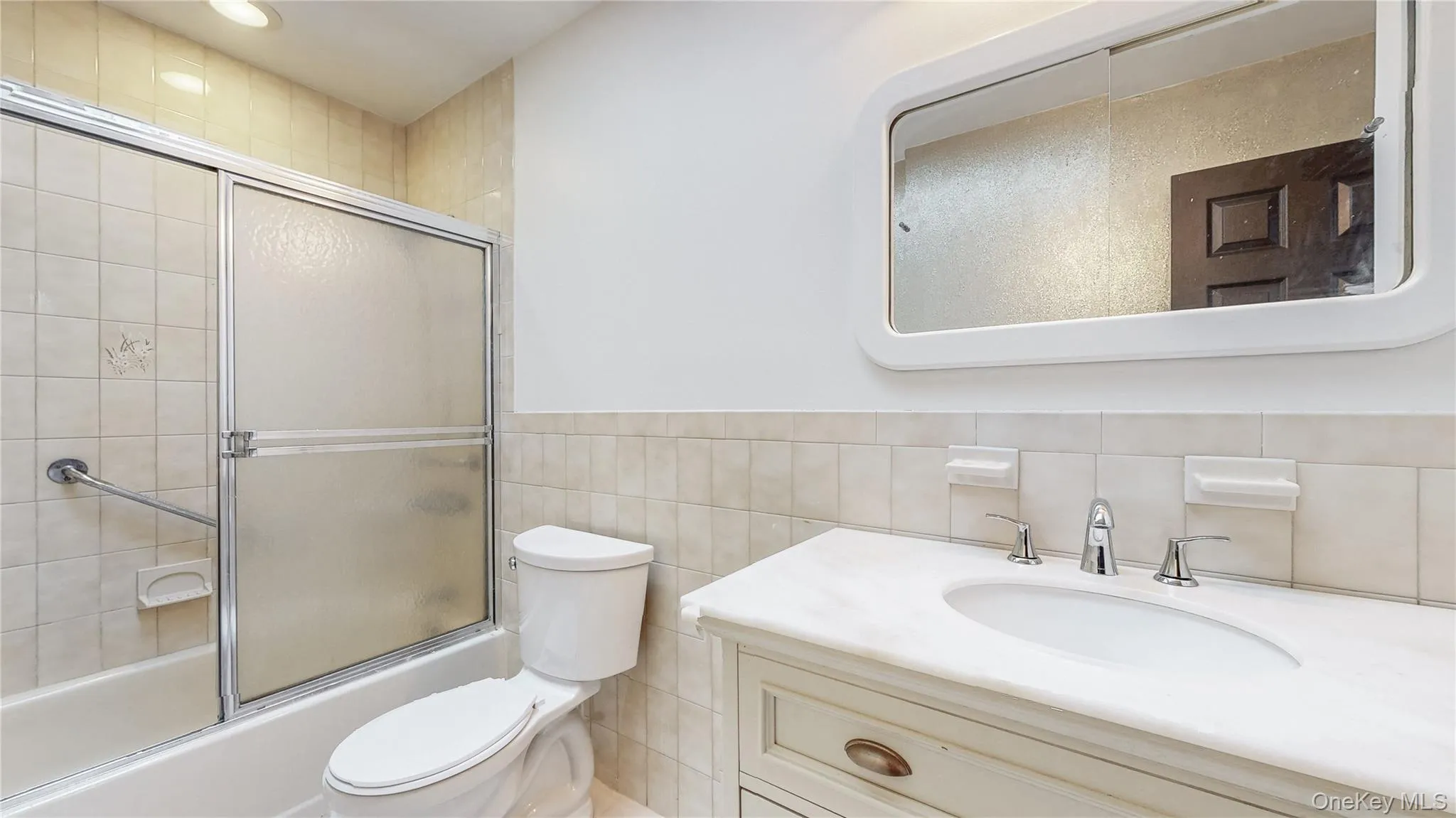 Full bathroom featuring tile walls, bath / shower combo with glass door, vanity, recessed lighting, and wainscoting Full bathroom featuring tile walls, bath / shower combo with glass door, vanity, recessed lighting, and wainscoting