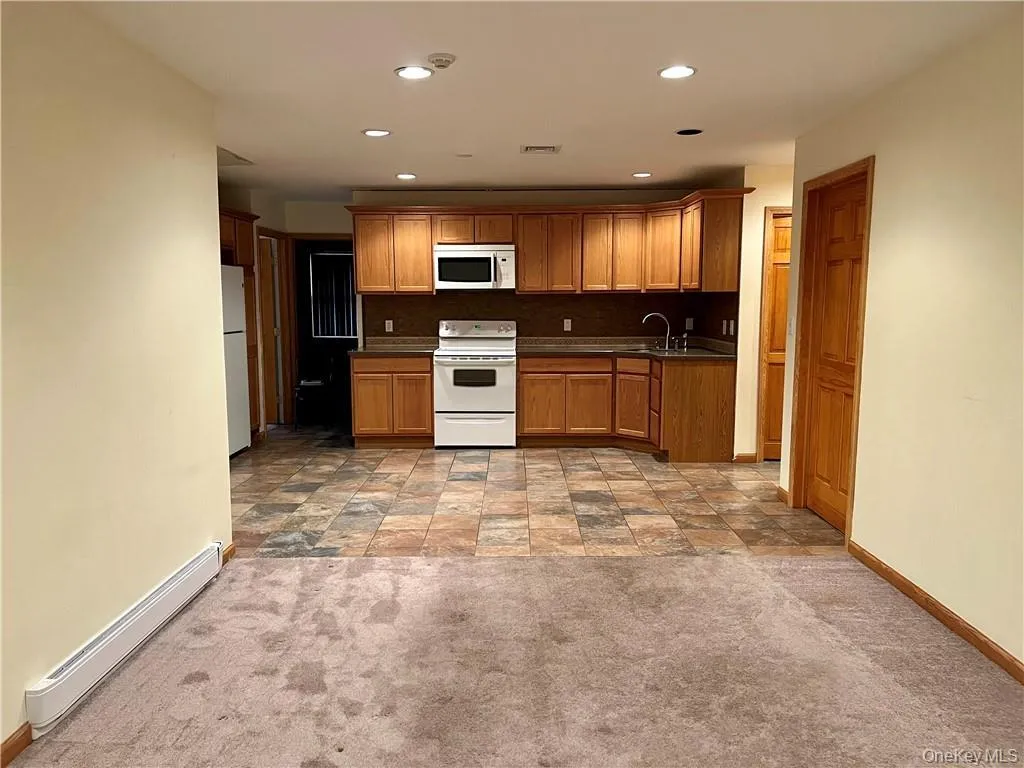 Kitchen featuring dark countertops, a baseboard radiator, white appliances, decorative backsplash, and light carpet Kitchen featuring dark countertops, a baseboard radiator, white appliances, decorative backsplash, and light carpet