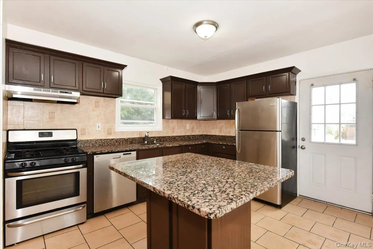 Kitchen featuring dark brown cabinetry, appliances with stainless steel finishes, light stone countertops, under cabinet range hood, and light tile patterned floors Kitchen featuring dark brown cabinetry, appliances with stainless steel finishes, light stone countertops, under cabinet range hood, and light tile patterned floors