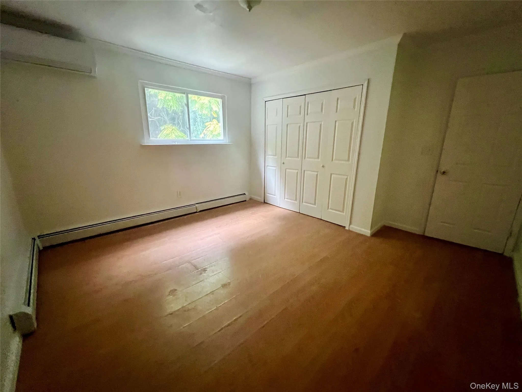 Unfurnished bedroom with crown molding, wood finished floors, a baseboard heating unit, a wall mounted air conditioner, and a closet Unfurnished bedroom with crown molding, wood finished floors, a baseboard heating unit, a wall mounted air conditioner, and a closet
