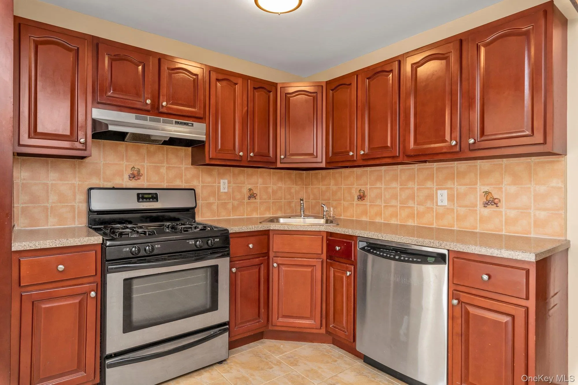 Kitchen featuring stainless steel appliances, tasteful backsplash, under cabinet range hood, light stone countertops, and light tile patterned flooring Kitchen featuring stainless steel appliances, tasteful backsplash, under cabinet range hood, light stone countertops, and light tile patterned flooring