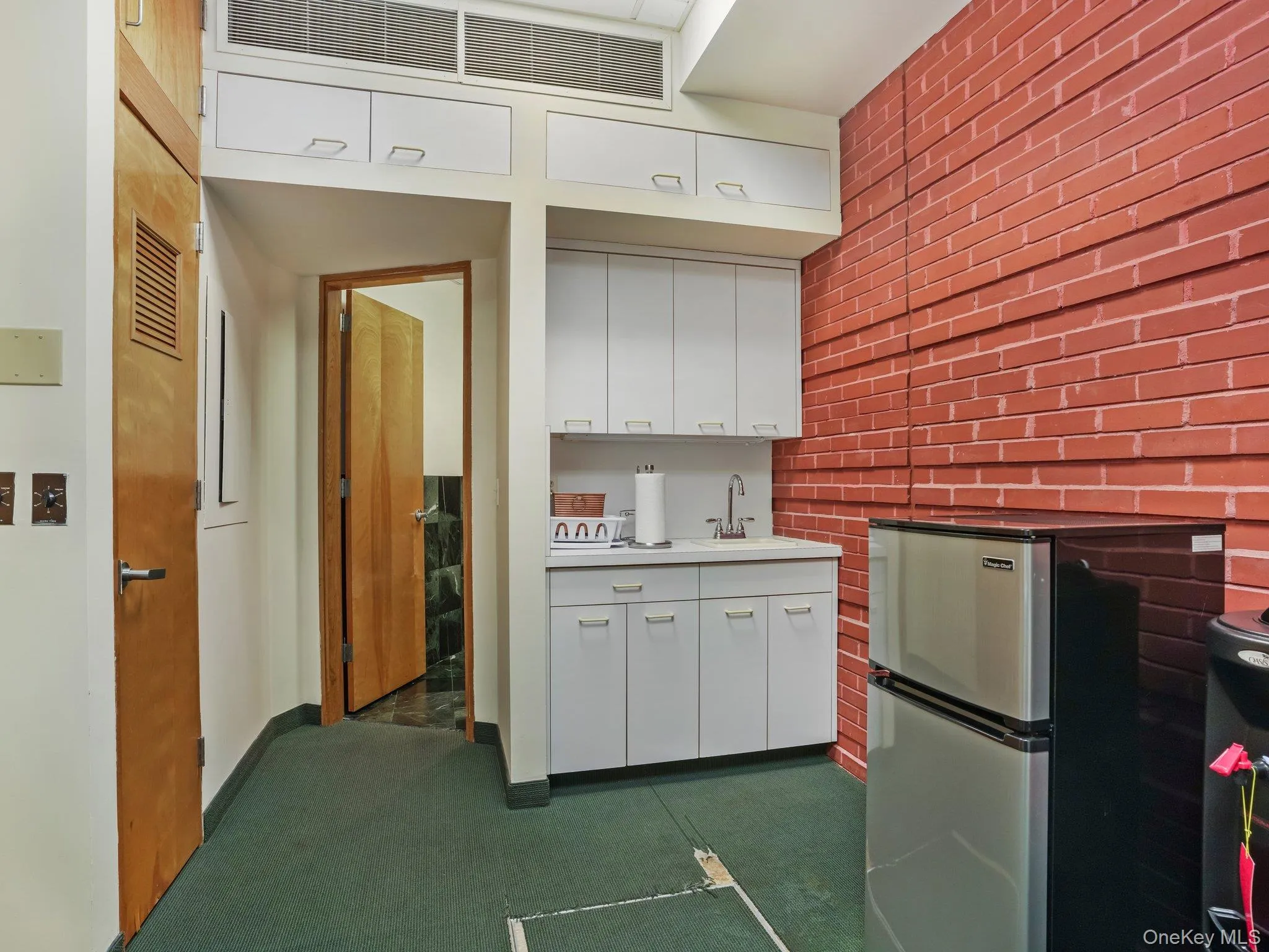 Kitchen with white cabinets, freestanding refrigerator, light countertops, brick wall, and dark carpet Kitchen with white cabinets, freestanding refrigerator, light countertops, brick wall, and dark carpet