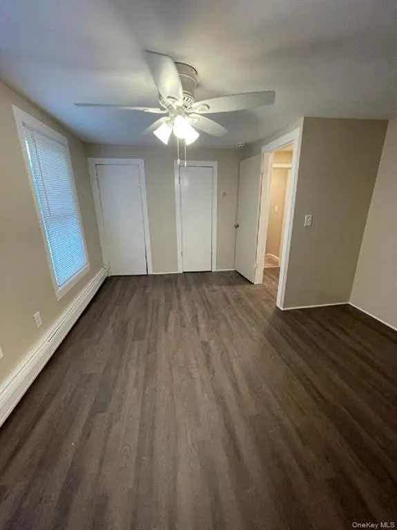 Unfurnished bedroom featuring a baseboard radiator, two closets, dark wood-style flooring, and a ceiling fan Unfurnished bedroom featuring a baseboard radiator, two closets, dark wood-style flooring, and a ceiling fan