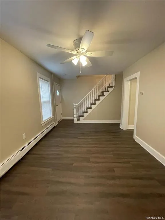 Unfurnished living room featuring stairs, a baseboard heating unit, dark wood-style floors, and a ceiling fan Unfurnished living room featuring stairs, a baseboard heating unit, dark wood-style floors, and a ceiling fan