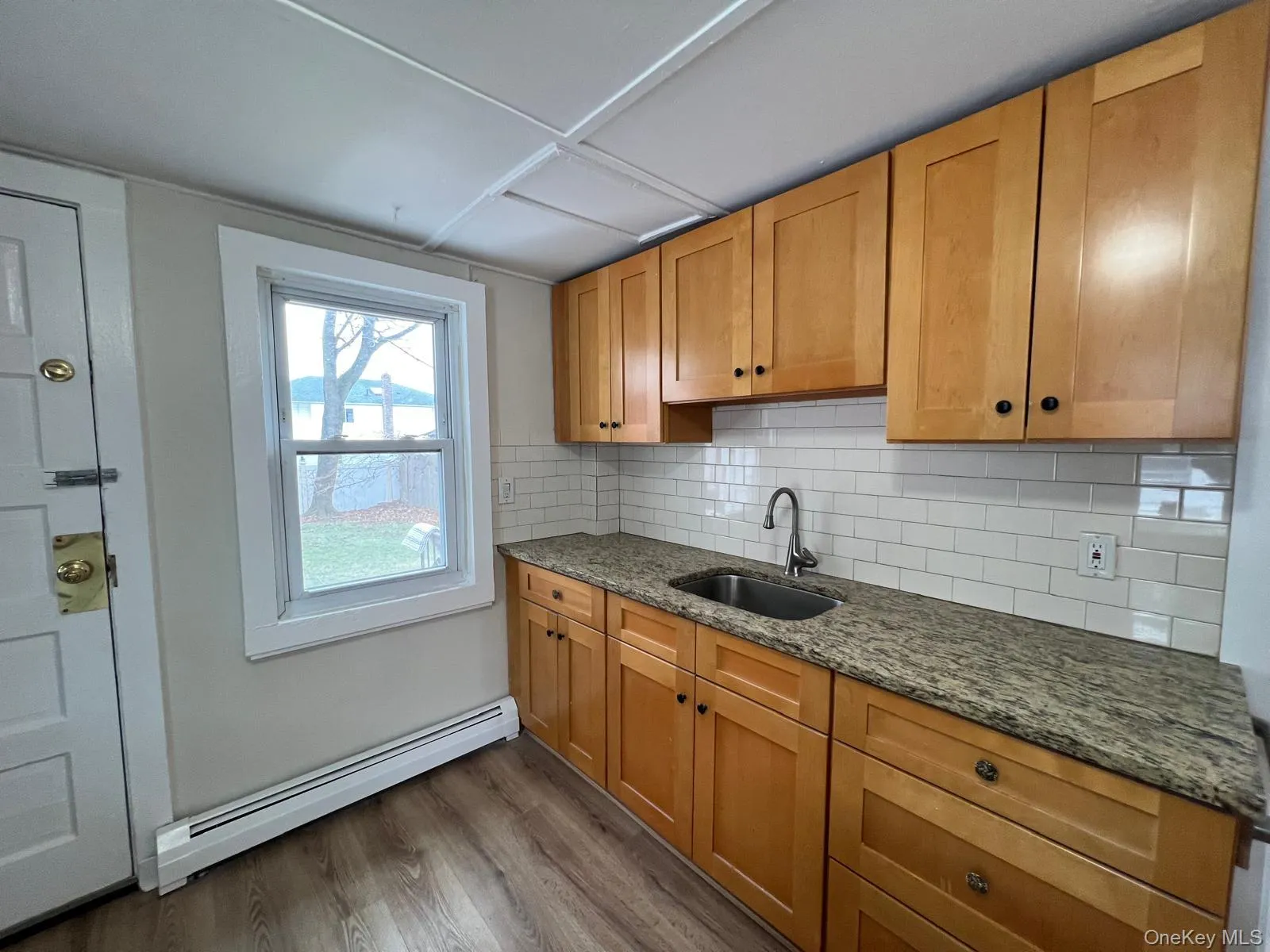 Kitchen featuring dark stone countertops, tasteful backsplash, a baseboard radiator, light wood-style flooring, and brown cabinets Kitchen featuring dark stone countertops, tasteful backsplash, a baseboard radiator, light wood-style flooring, and brown cabinets