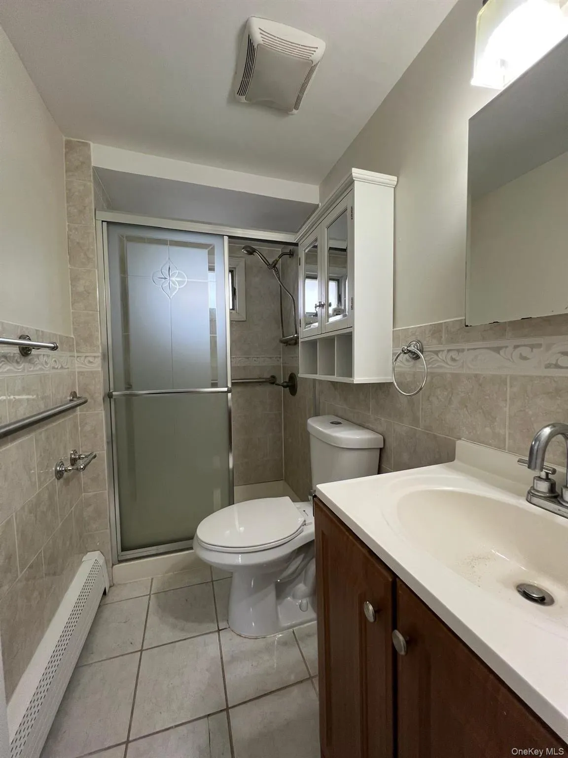 Full bath featuring tile walls, a baseboard heating unit, vanity, a stall shower, and light tile patterned flooring Full bath featuring tile walls, a baseboard heating unit, vanity, a stall shower, and light tile patterned flooring
