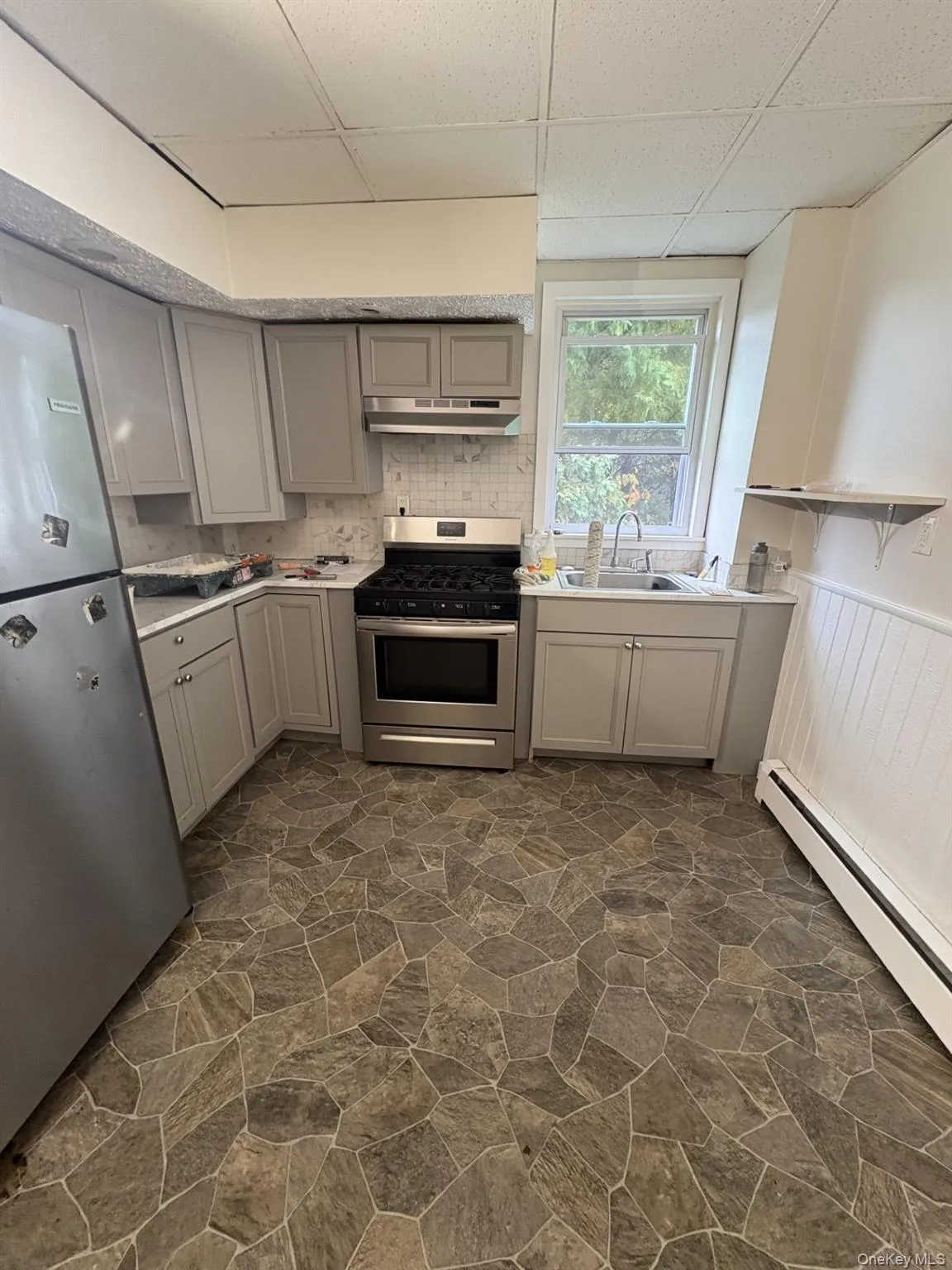 Kitchen featuring a baseboard heating unit, stainless steel appliances, gray cabinetry, a drop ceiling, and tasteful backsplash Kitchen featuring a baseboard heating unit, stainless steel appliances, gray cabinetry, a drop ceiling, and tasteful backsplash