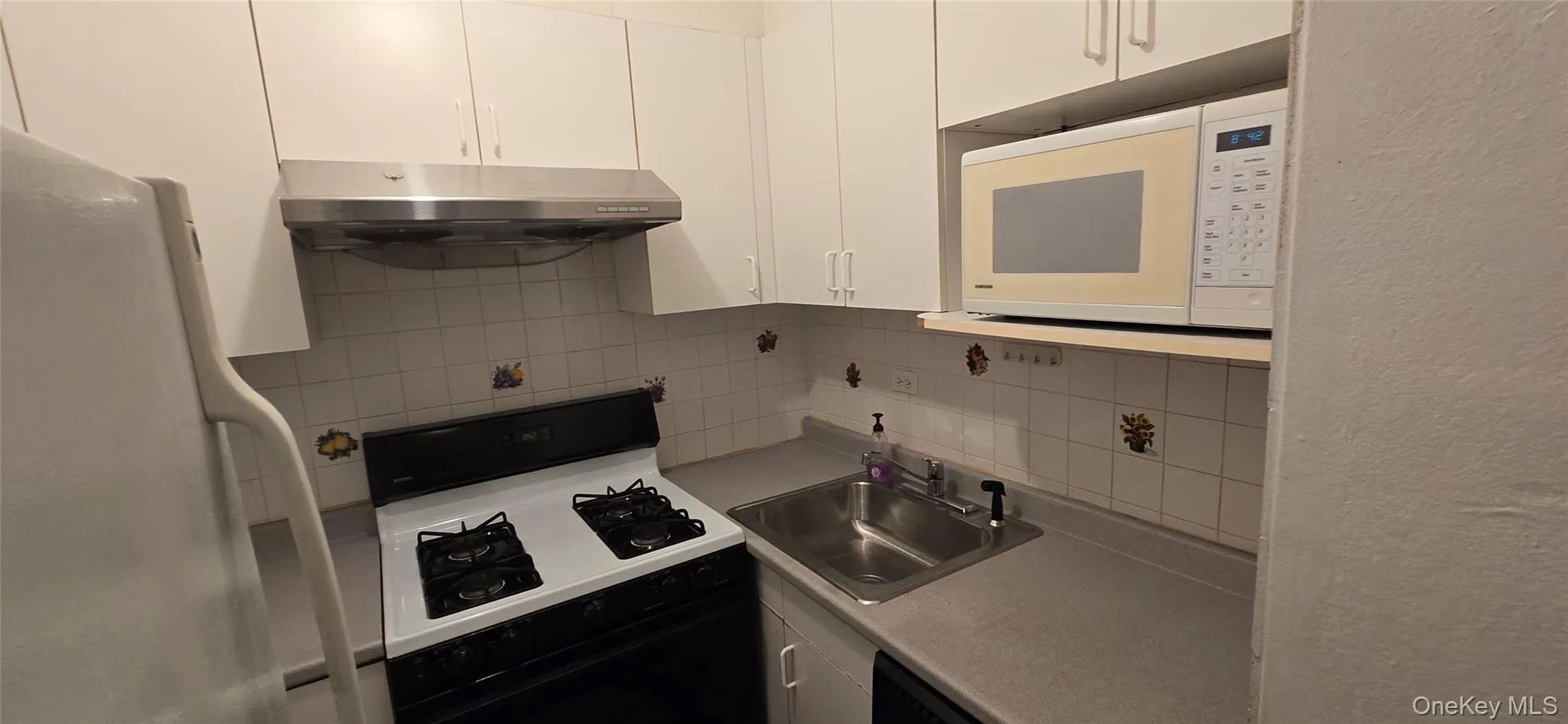 Kitchen featuring tasteful backsplash, white appliances, under cabinet range hood, light countertops, and white cabinetry Kitchen featuring tasteful backsplash, white appliances, under cabinet range hood, light countertops, and white cabinetry