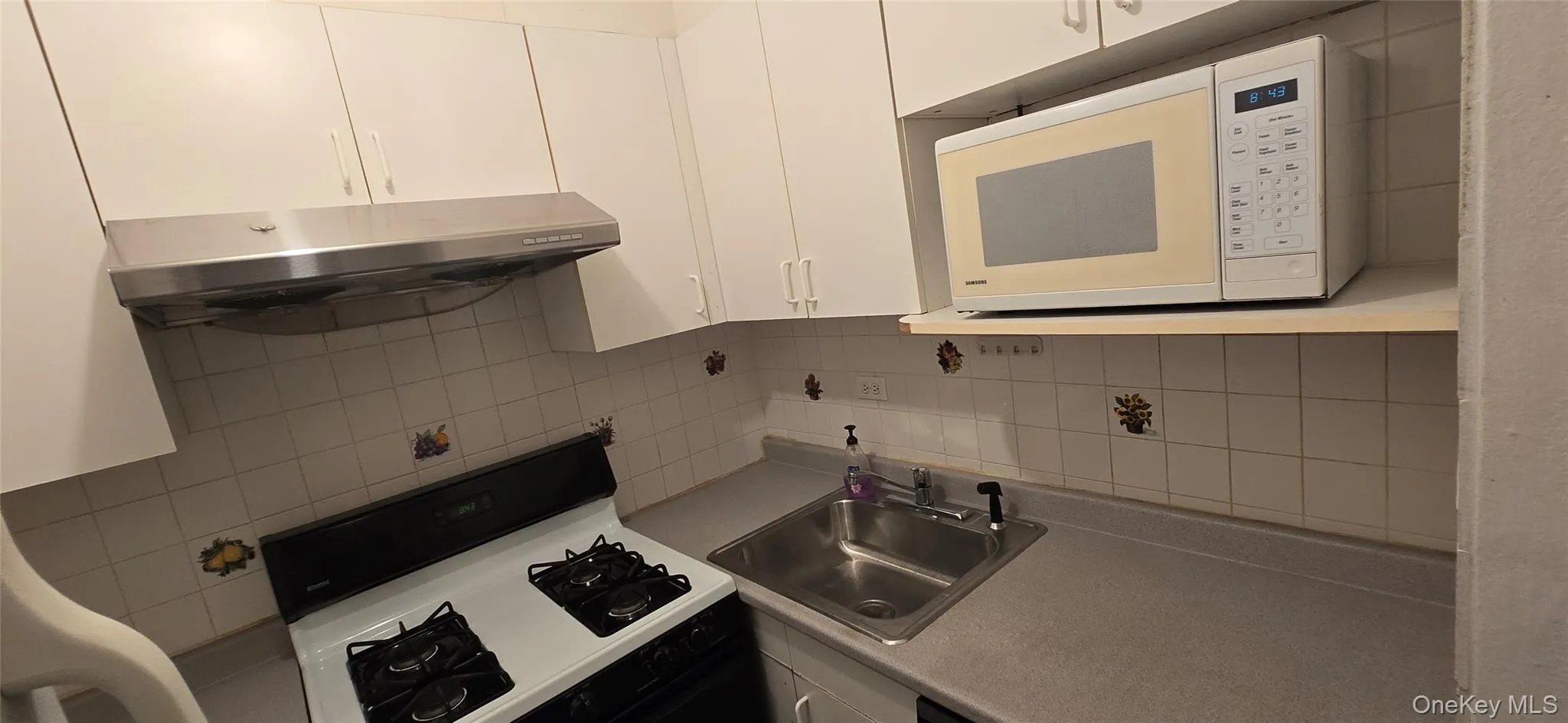 Kitchen featuring black gas range oven, backsplash, white microwave, range hood, and white cabinetry Kitchen featuring black gas range oven, backsplash, white microwave, range hood, and white cabinetry