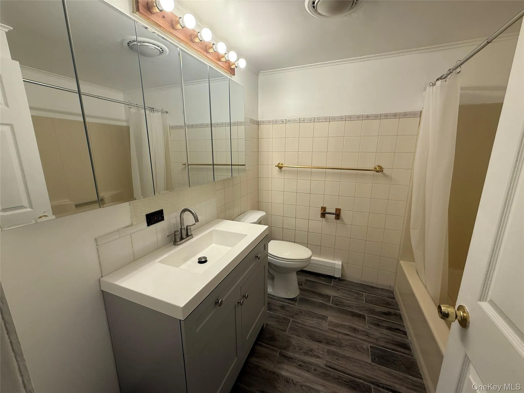 Full bath featuring tile walls, wood finish floors, vanity, shower / bath combo with shower curtain, and baseboard heating Full bath featuring tile walls, wood finish floors, vanity, shower / bath combo with shower curtain, and baseboard heating