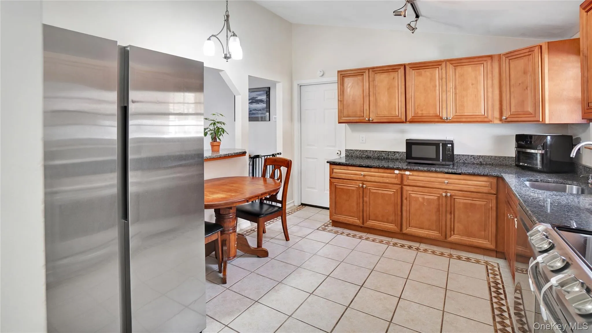 Kitchen featuring stainless steel appliances, light tile patterned flooring, dark stone counters, brown cabinets, and decorative light fixtures Kitchen featuring stainless steel appliances, light tile patterned flooring, dark stone counters, brown cabinets, and decorative light fixtures