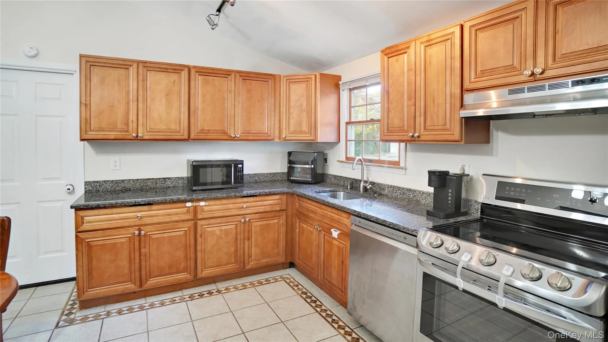 Kitchen featuring stainless steel appliances, inlaid floor details, light tile patterned floors, dark stone countertops, and under cabinet range hood Kitchen featuring stainless steel appliances, inlaid floor details, light tile patterned floors, dark stone countertops, and under cabinet range hood