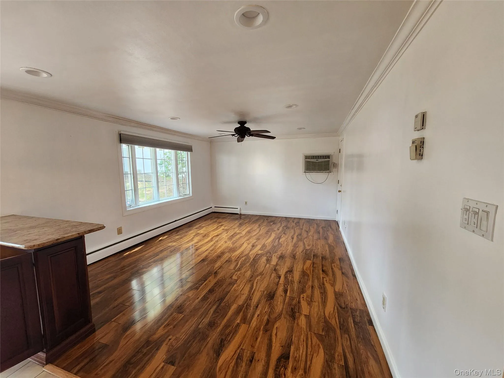 Unfurnished living room with crown molding, dark wood-style flooring, a baseboard heating unit, ceiling fan, and a wall unit AC Unfurnished living room with crown molding, dark wood-style flooring, a baseboard heating unit, ceiling fan, and a wall unit AC