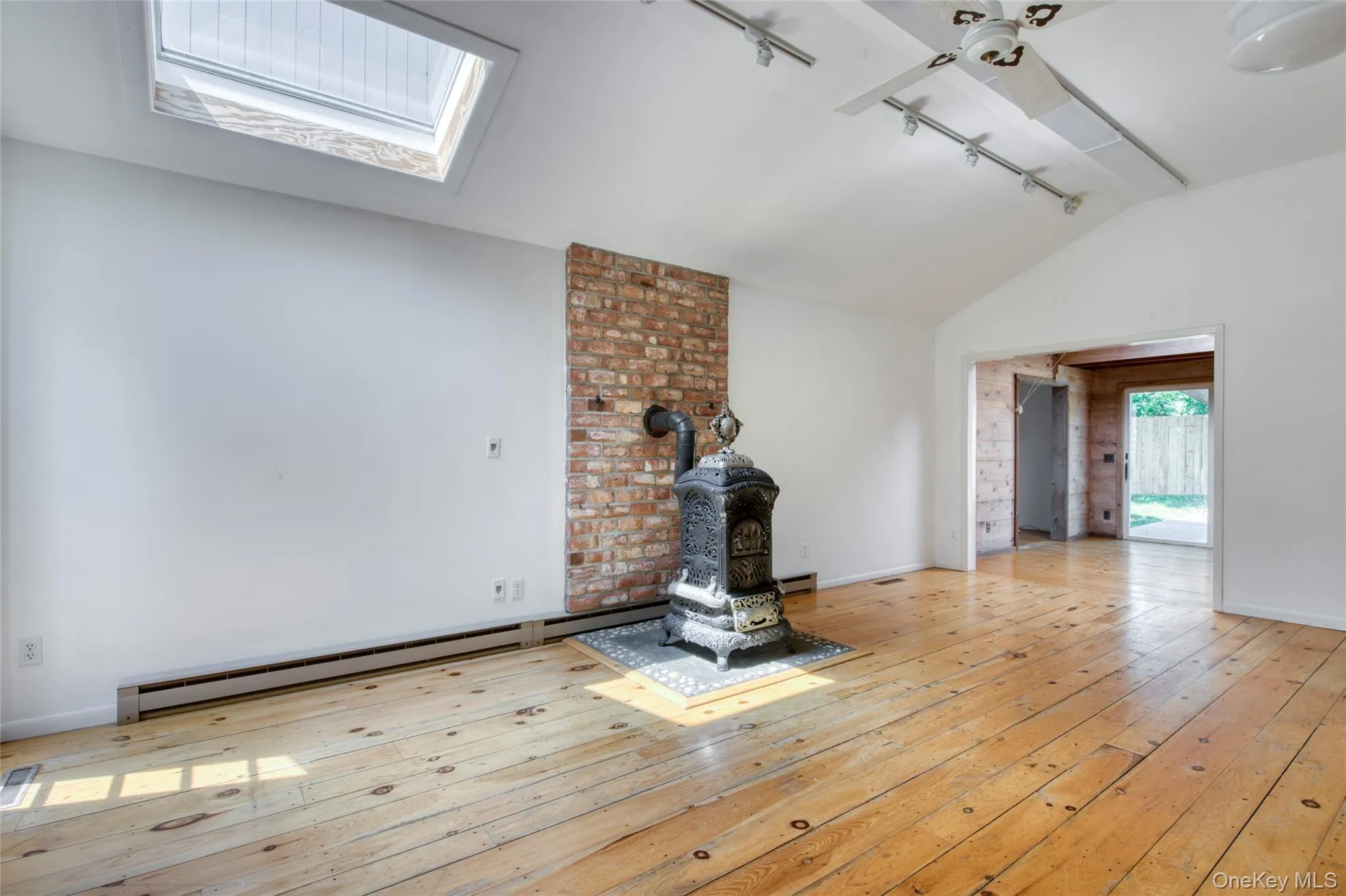 Unfurnished living room with a wood stove, track lighting, light wood finished floors, lofted ceiling, and a baseboard radiator Unfurnished living room with a wood stove, track lighting, light wood finished floors, lofted ceiling, and a baseboard radiator