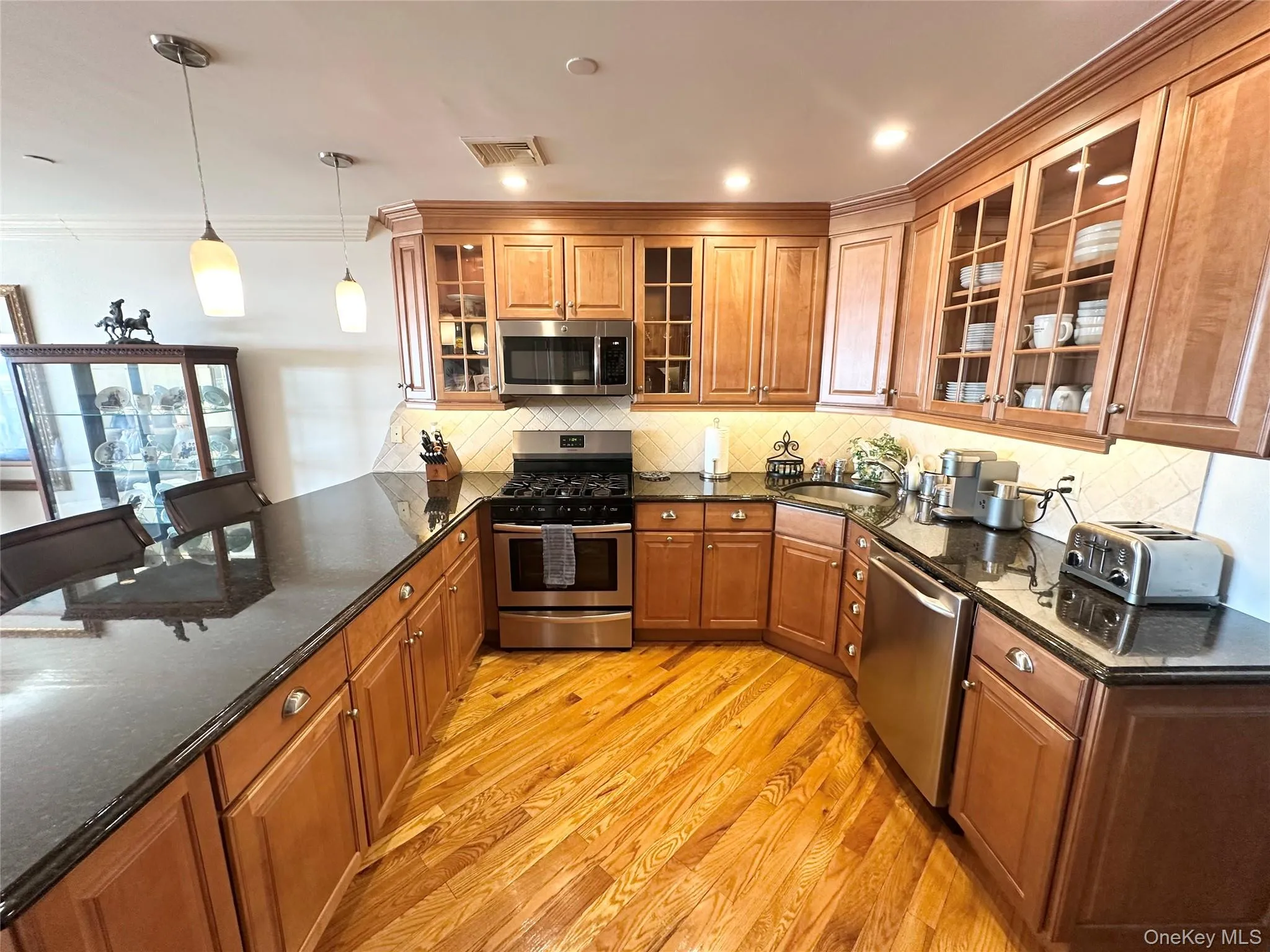 Kitchen with brown cabinetry, appliances with stainless steel finishes, glass insert cabinets, tasteful backsplash, and dark stone countertops Kitchen with brown cabinetry, appliances with stainless steel finishes, glass insert cabinets, tasteful backsplash, and dark stone countertops