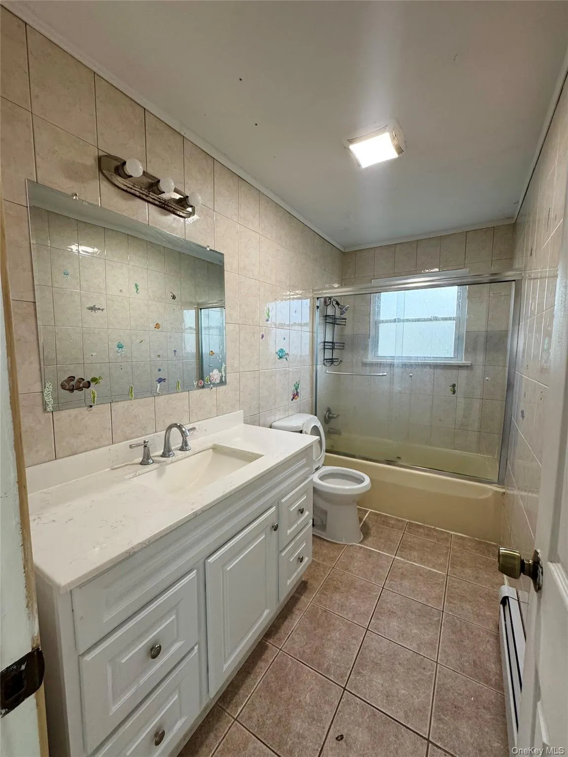 Full bathroom featuring tile walls, combined bath / shower with glass door, vanity, light tile patterned floors, and baseboard heating Full bathroom featuring tile walls, combined bath / shower with glass door, vanity, light tile patterned floors, and baseboard heating