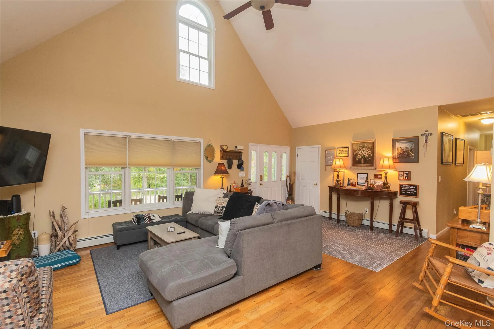 Living room featuring high vaulted ceiling, plenty of natural light, light wood finished floors, a ceiling fan, and a baseboard heating unit Living room featuring high vaulted ceiling, plenty of natural light, light wood finished floors, a ceiling fan, and a baseboard heating unit