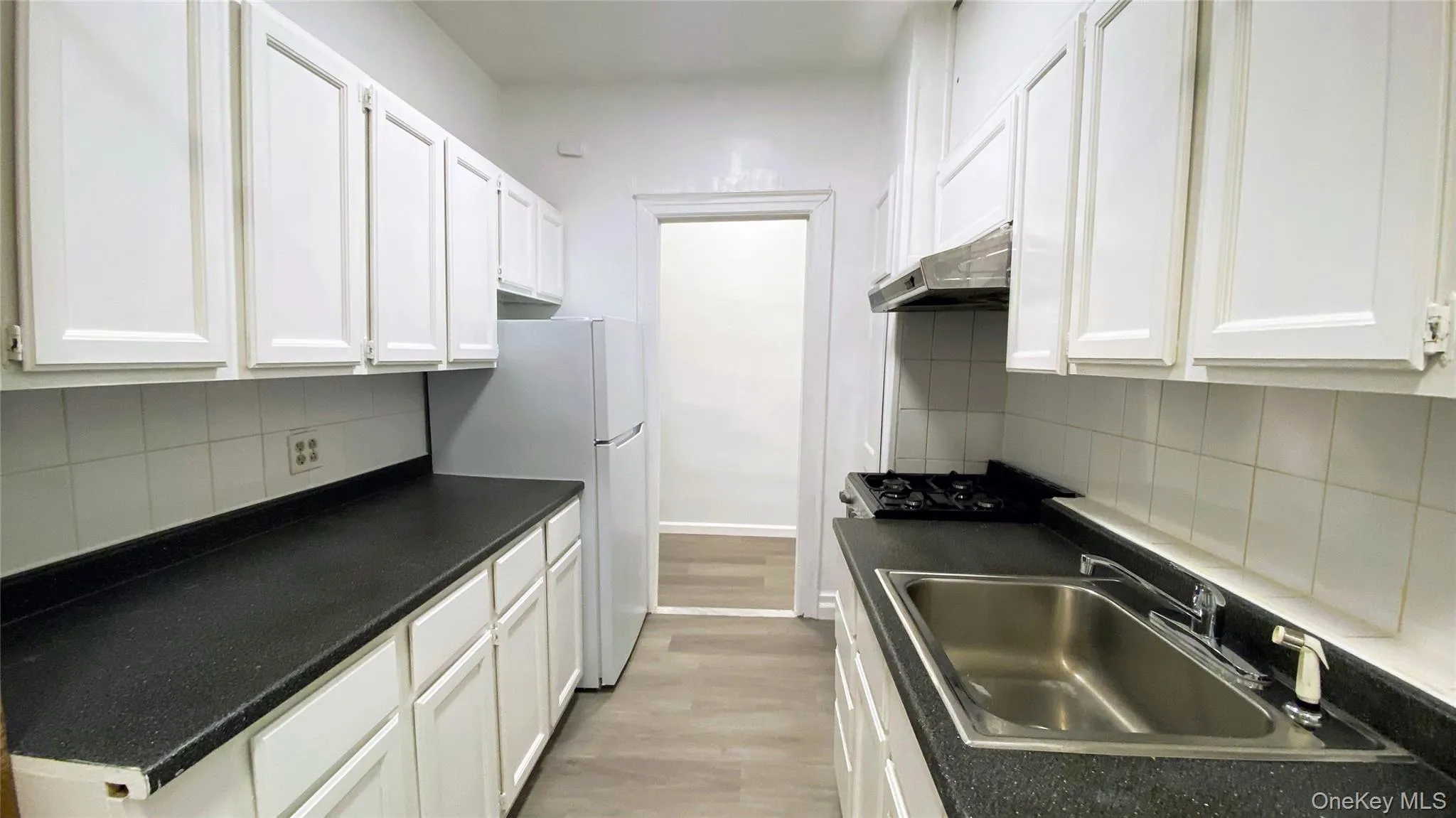 Kitchen with decorative backsplash, white cabinets, dark countertops, freestanding refrigerator, and under cabinet range hood Kitchen with decorative backsplash, white cabinets, dark countertops, freestanding refrigerator, and under cabinet range hood