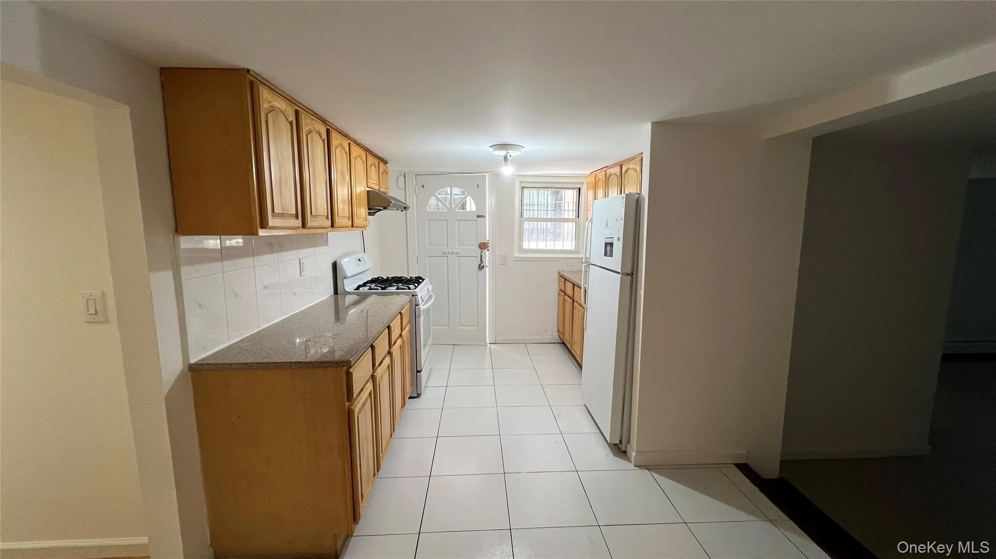 Kitchen featuring white appliances, dark stone counters, light tile patterned floors, backsplash, and brown cabinetry Kitchen featuring white appliances, dark stone counters, light tile patterned floors, backsplash, and brown cabinetry