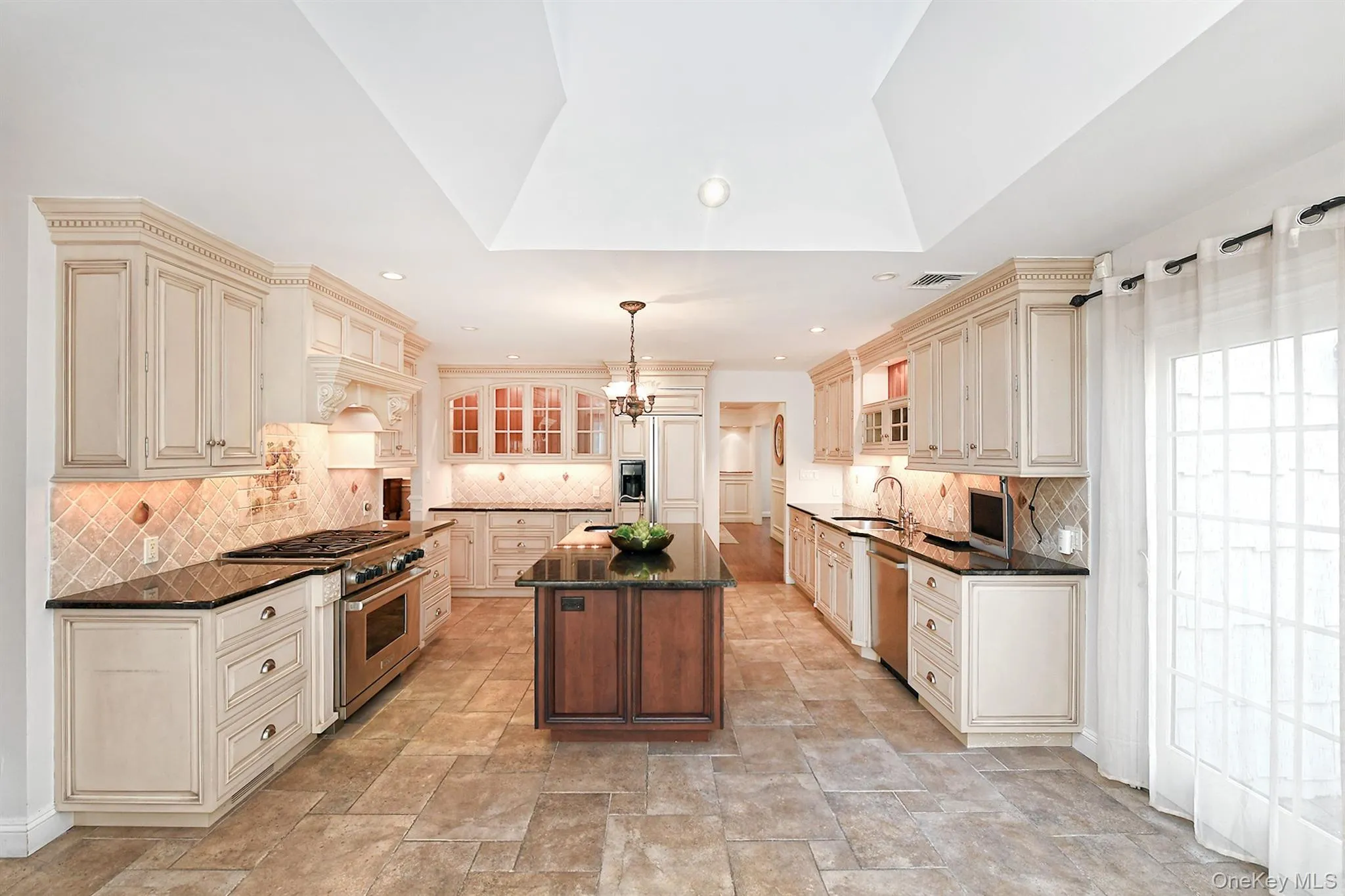 Kitchen with cream cabinetry, glass insert cabinets, a center island, dark stone counters, and appliances with stainless steel finishes Kitchen with cream cabinetry, glass insert cabinets, a center island, dark stone counters, and appliances with stainless steel finishes