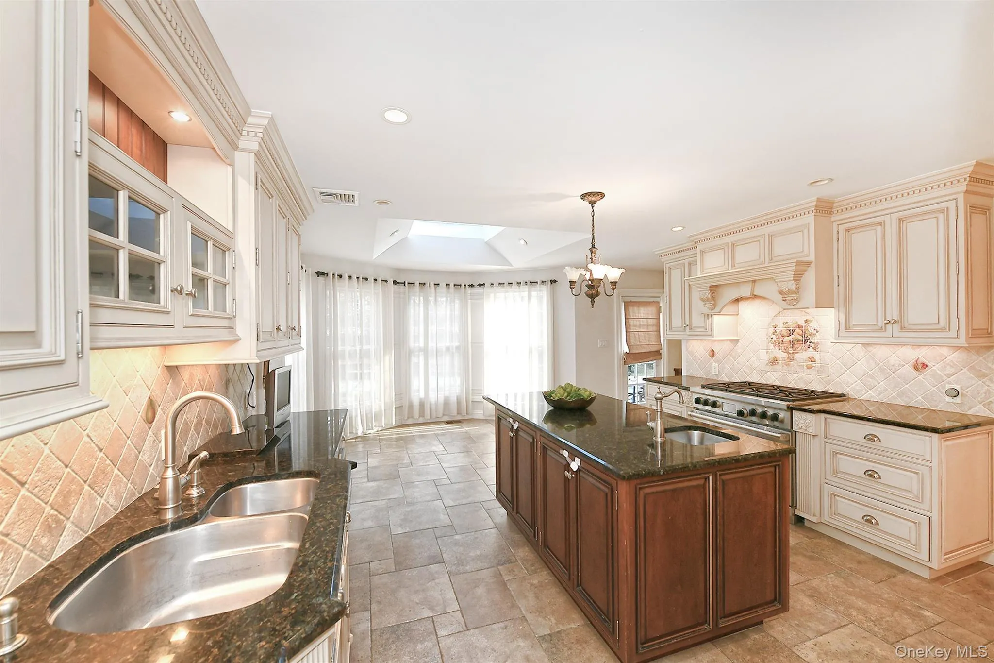 Kitchen with decorative backsplash, cream cabinetry, a kitchen island with sink, a skylight, and a tray ceiling Kitchen with decorative backsplash, cream cabinetry, a kitchen island with sink, a skylight, and a tray ceiling