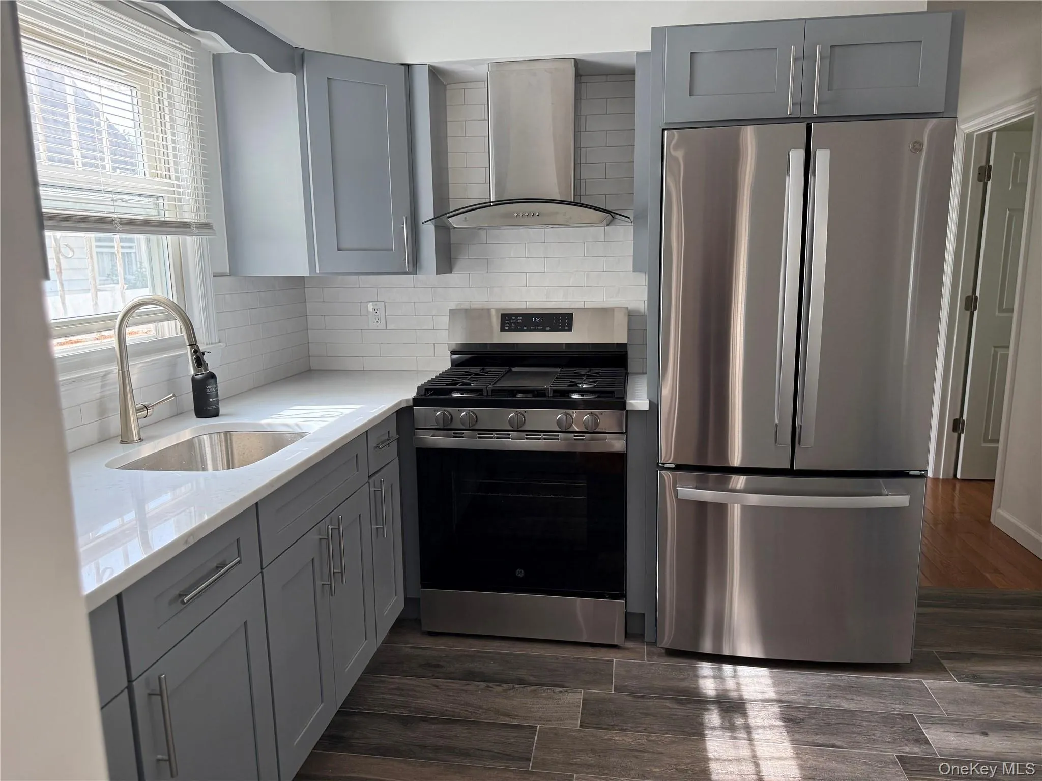 Kitchen with gray cabinetry, appliances with stainless steel finishes, wall chimney range hood, decorative backsplash, and light stone countertops Kitchen with gray cabinetry, appliances with stainless steel finishes, wall chimney range hood, decorative backsplash, and light stone countertops