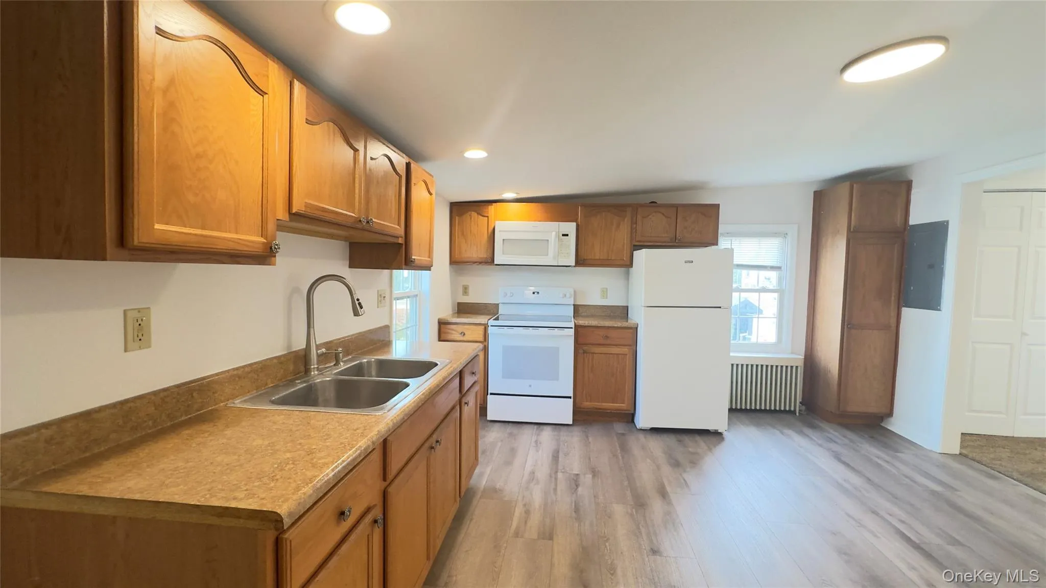 Kitchen featuring white appliances, recessed lighting, brown cabinetry, light wood-style flooring, and radiator heating unit Kitchen featuring white appliances, recessed lighting, brown cabinetry, light wood-style flooring, and radiator heating unit