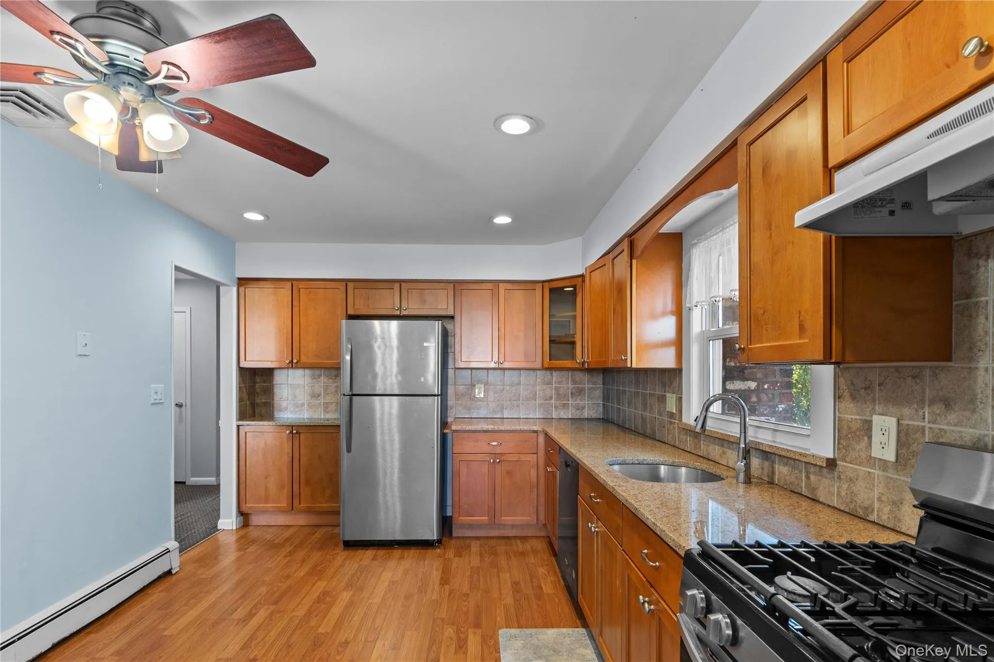 Kitchen featuring stainless steel appliances, light stone counters, brown cabinetry, a baseboard radiator, and backsplash Kitchen featuring stainless steel appliances, light stone counters, brown cabinetry, a baseboard radiator, and backsplash