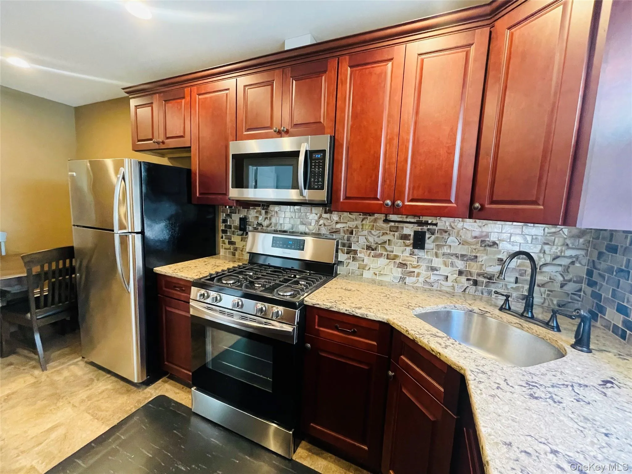Kitchen featuring decorative backsplash, appliances with stainless steel finishes, light stone countertops, and reddish brown cabinets Kitchen featuring decorative backsplash, appliances with stainless steel finishes, light stone countertops, and reddish brown cabinets